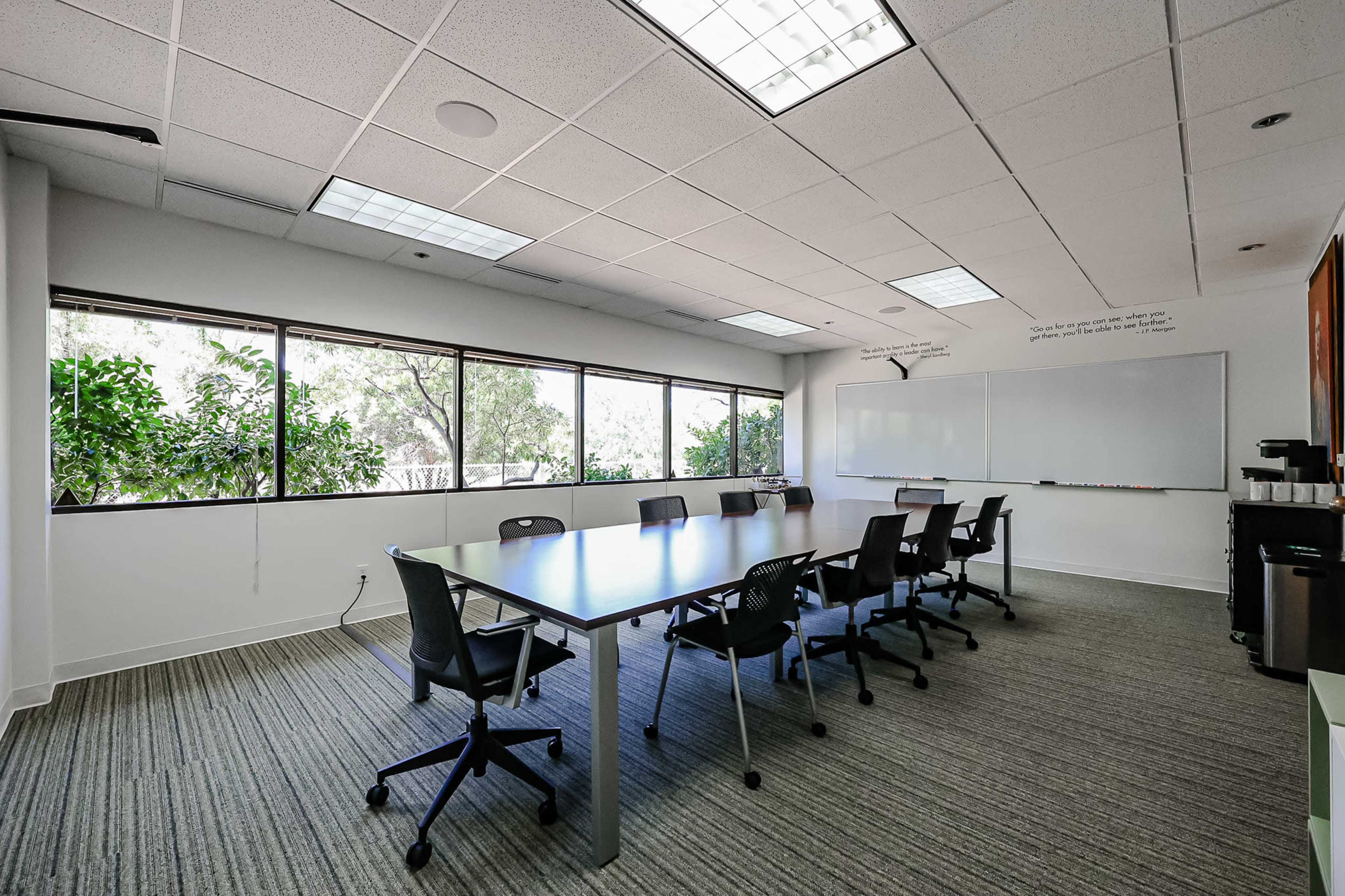 A conference room features a long table surrounded by black chairs, with large windows allowing natural light and greenery visible outside.