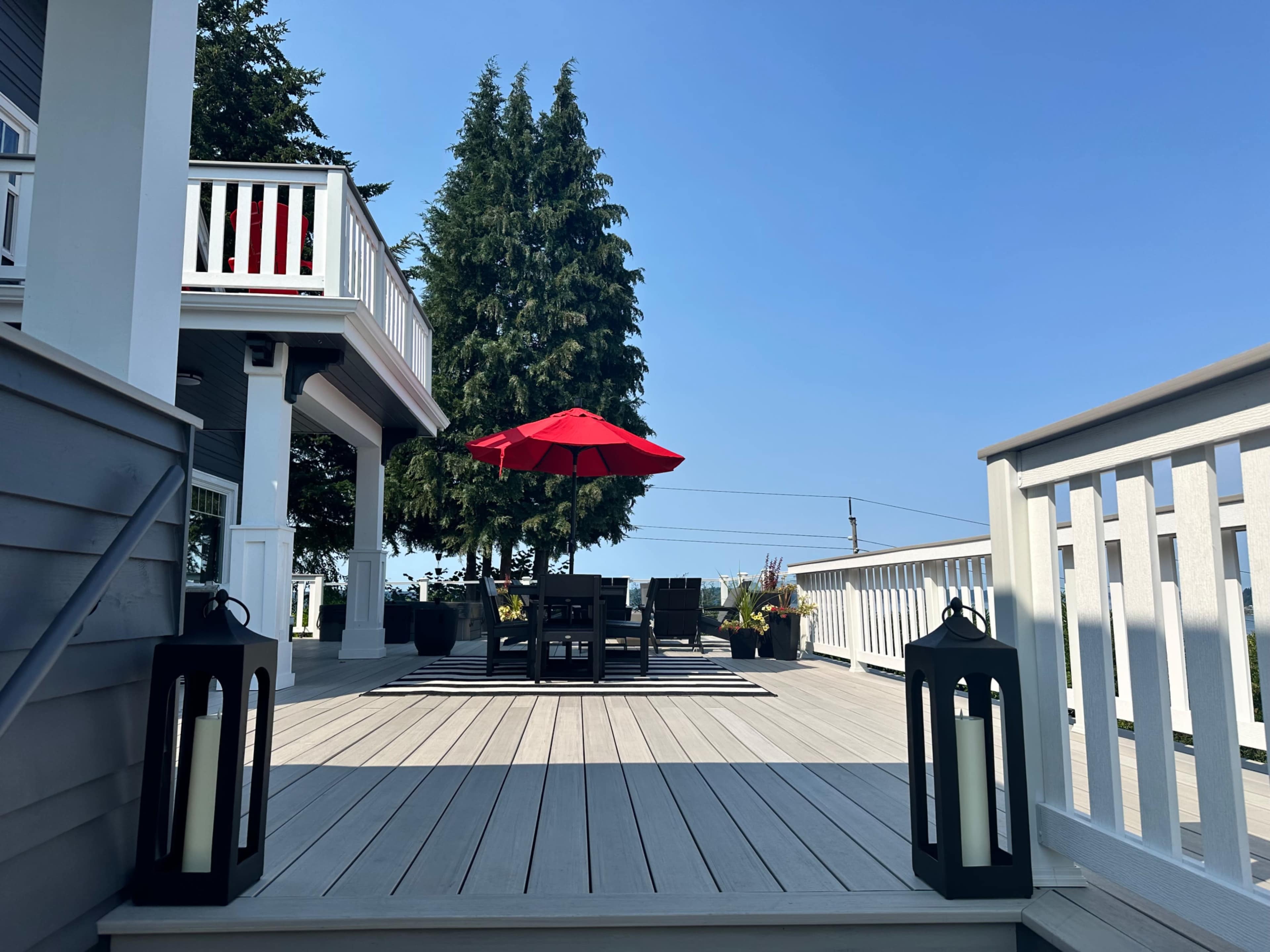 The image shows a deck area with a red umbrella over a seating arrangement, two lanterns placed on the steps, and a house with a railing in the background.