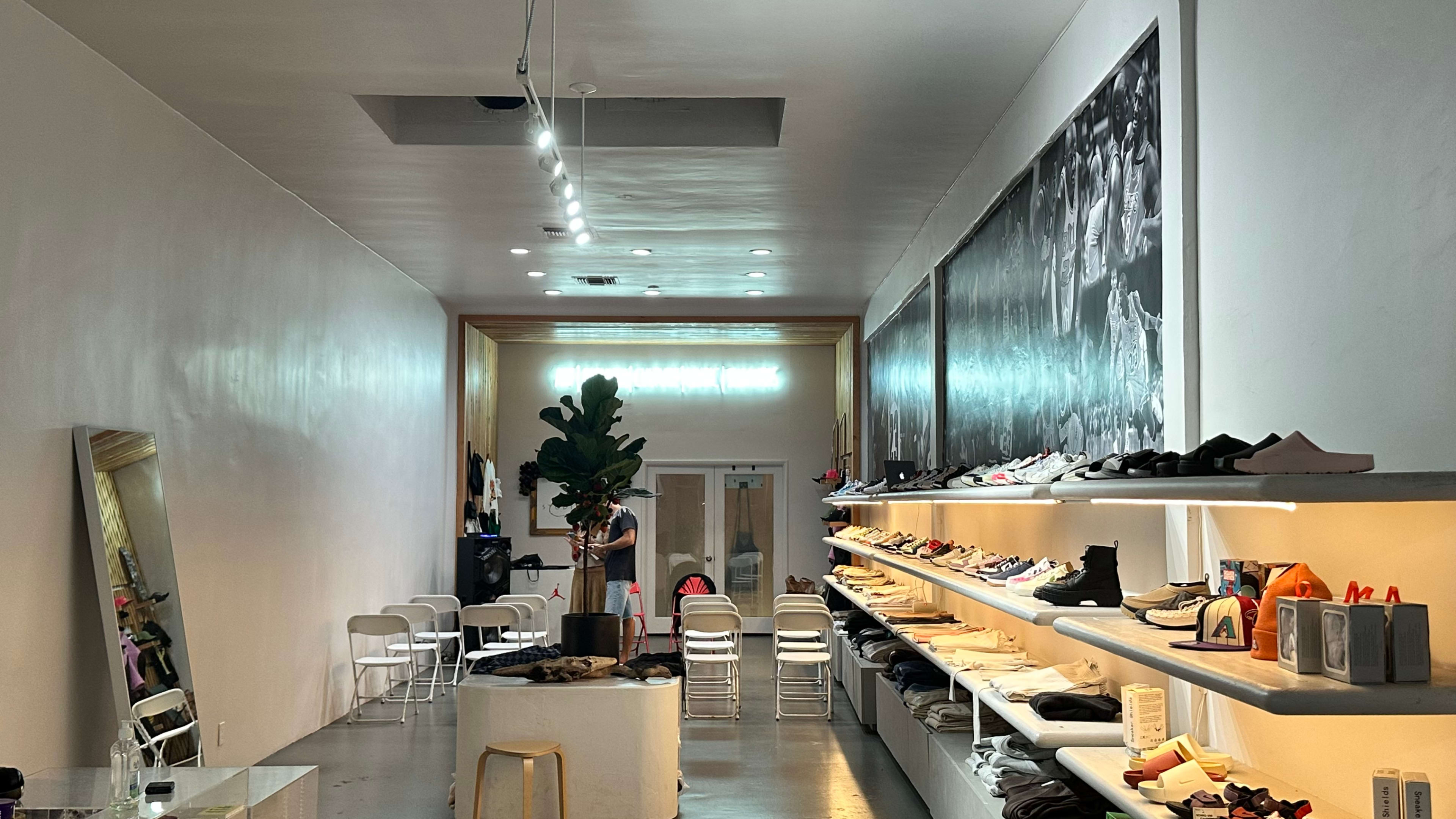 The image shows a contemporary shoe store with minimalist design, featuring shelves lined with various footwear along the walls and seating arrangements in the center.