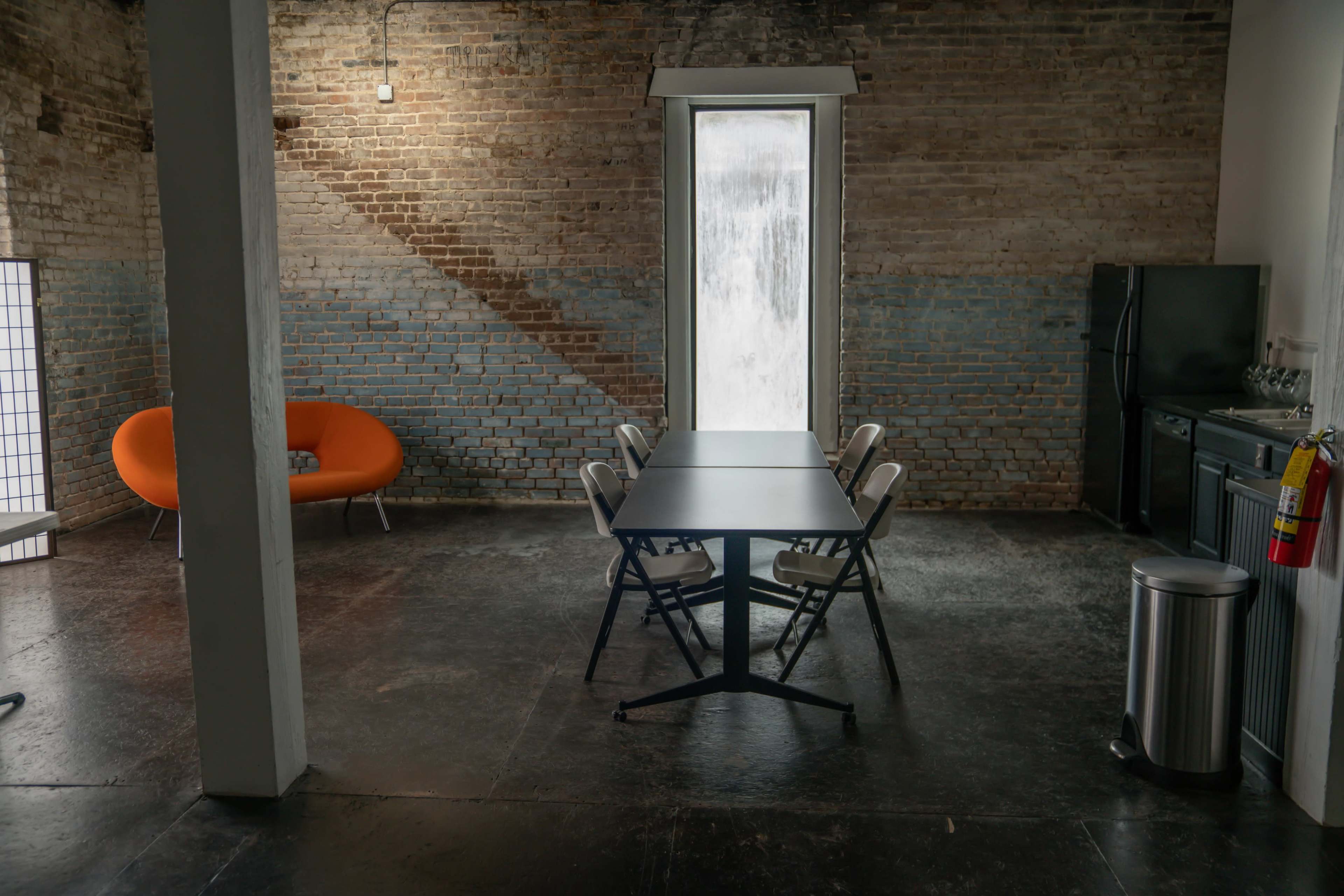 A meeting space features a rectangular table surrounded by chairs, an orange couch, and a large window with a view of a brick wall.