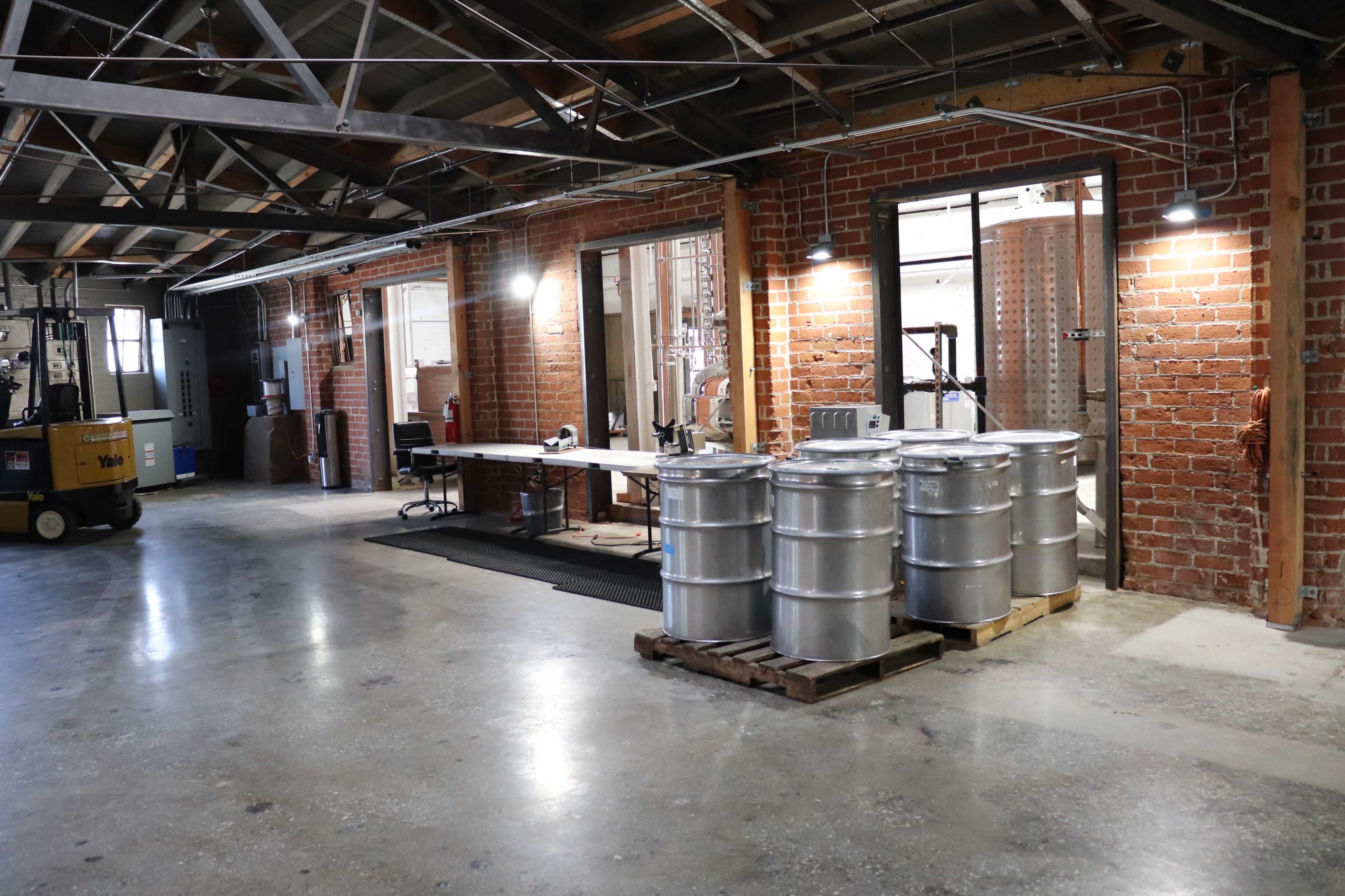 An industrial interior with silver barrels on a wooden pallet, a work table, and a forklift in a brick building.