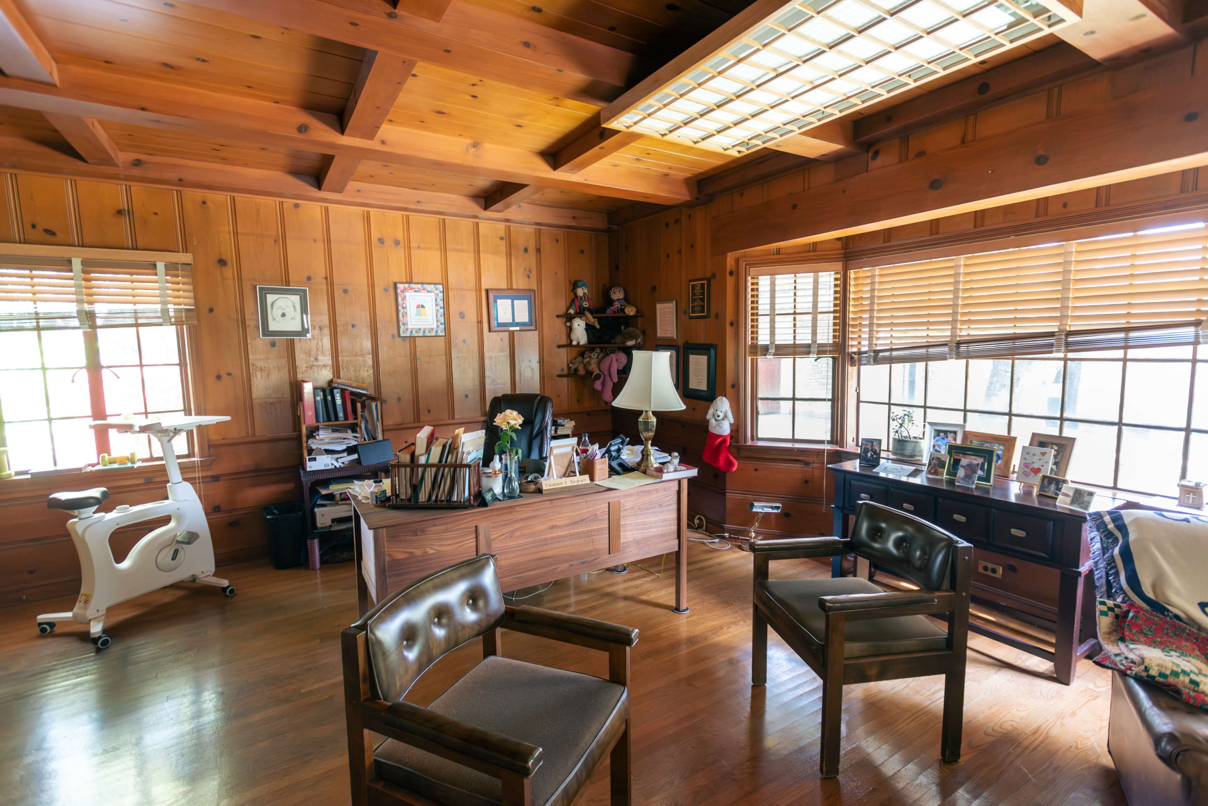 The office features wooden walls, a large desk with various items, and several chairs arranged around the space.