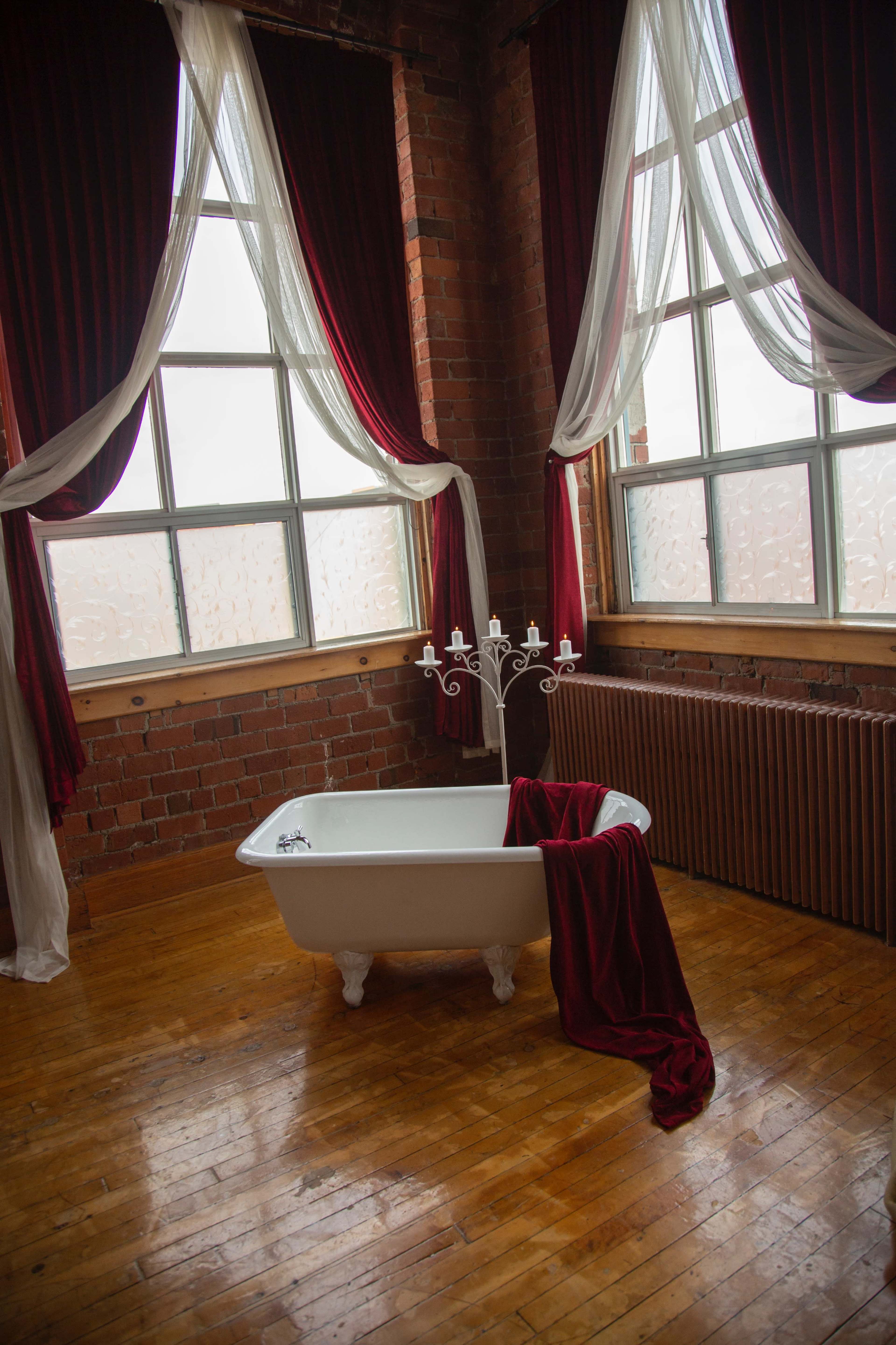 A freestanding white bathtub with a red draped cloth sits on a wooden floor beneath large windows framed by red curtains.