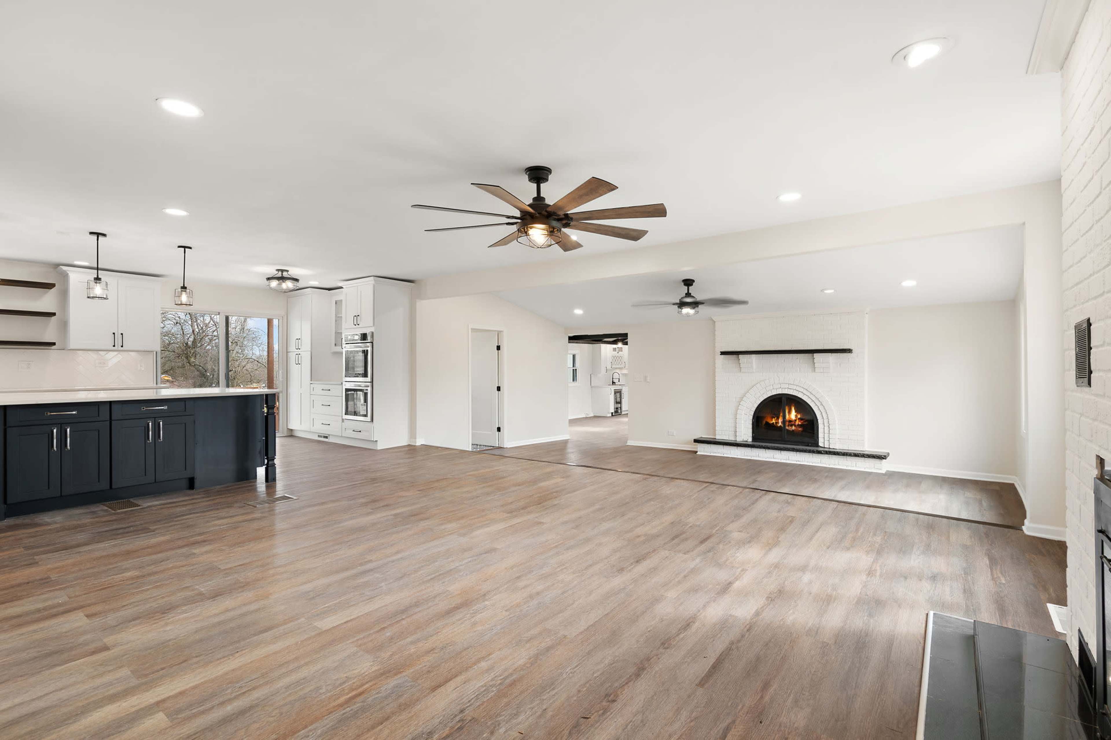 The image shows an open-concept living space featuring a kitchen with dark cabinets, a bright area with large windows, and a fireplace in the adjacent living room.