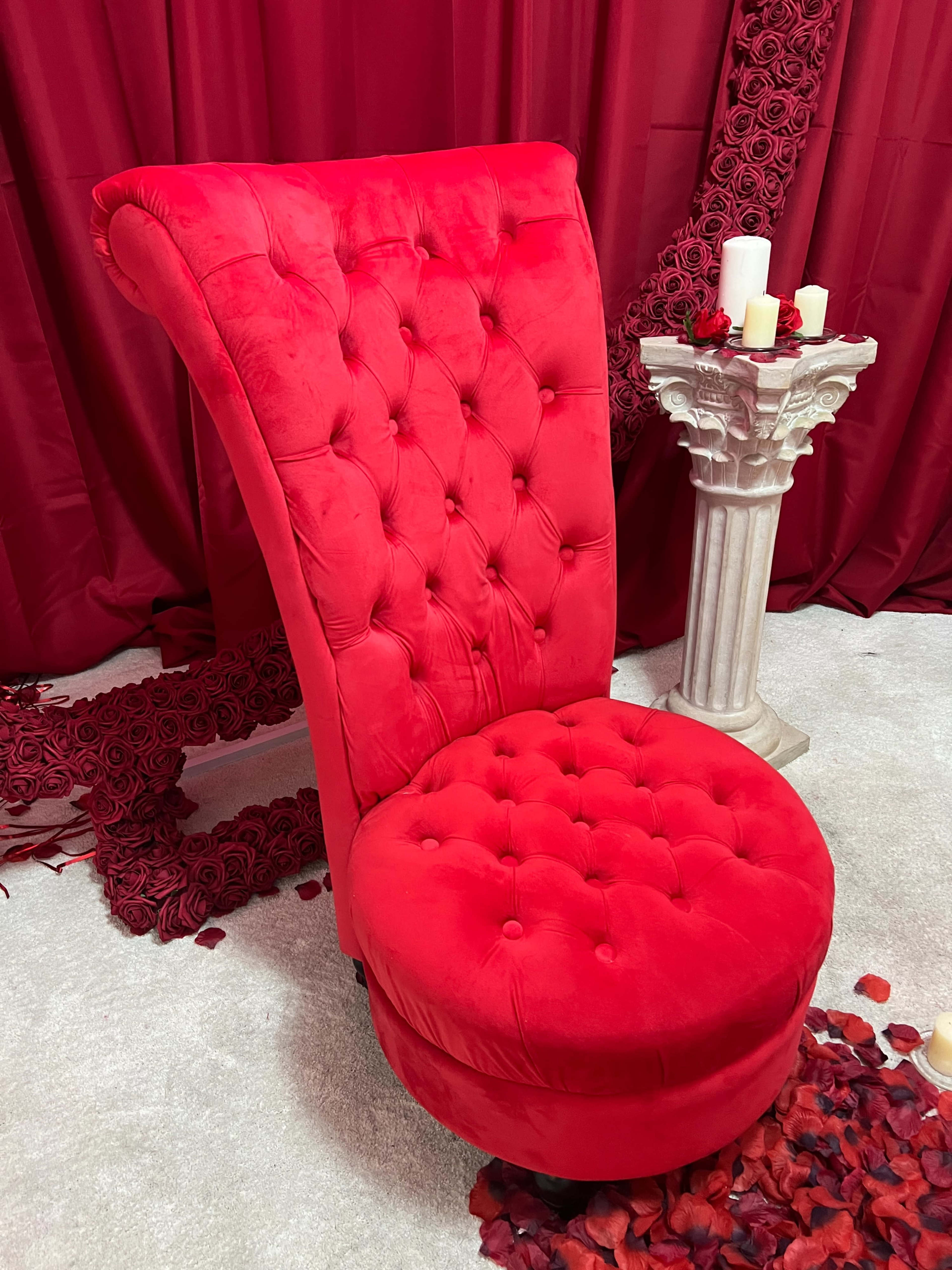 A red, tufted chaise lounge positioned alongside a column with candles, set against a backdrop of red fabric and rose petals.
