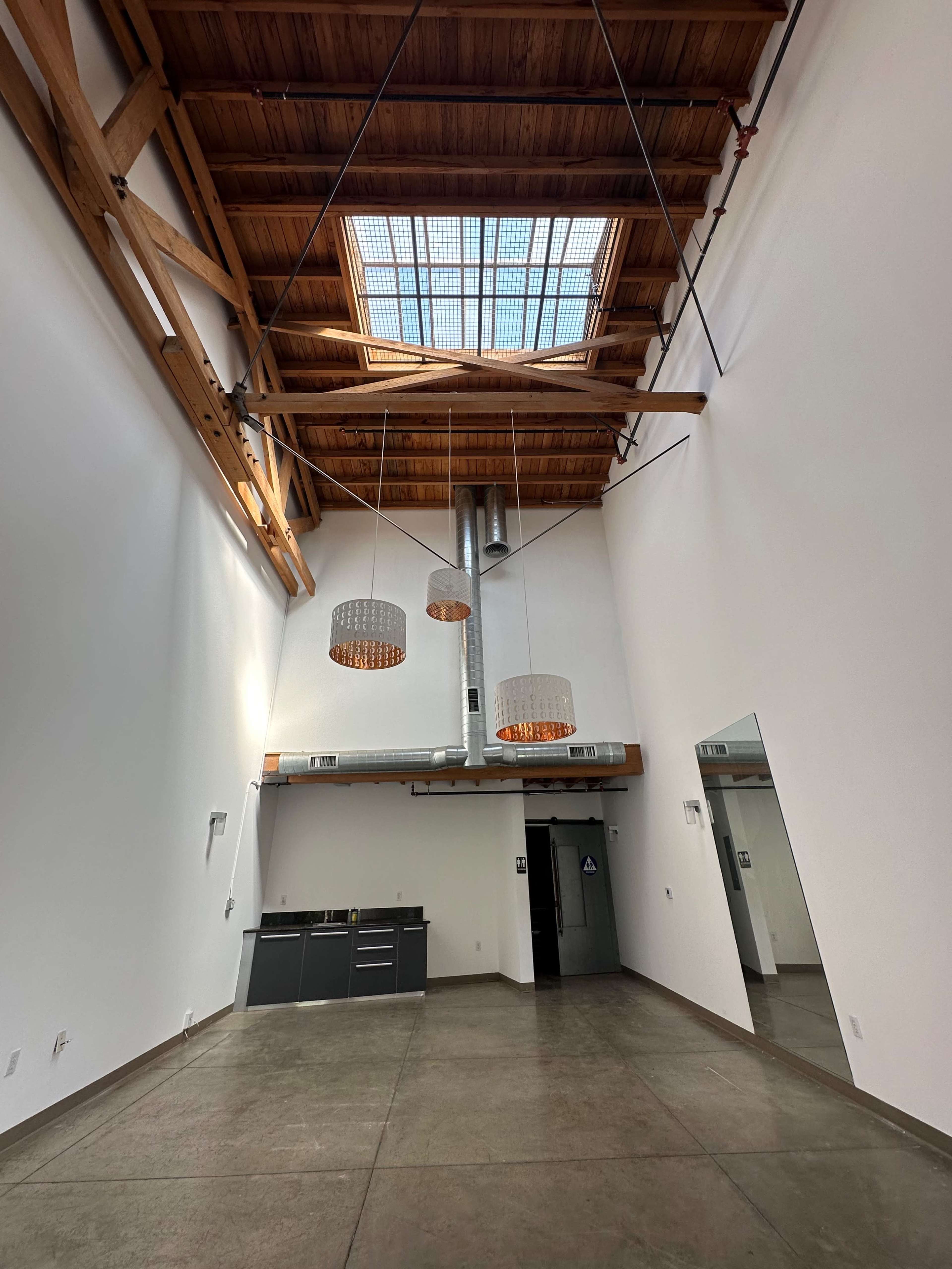 The image shows a spacious interior with a high wooden ceiling, large skylight, and modern light fixtures, featuring a kitchen area along one wall.