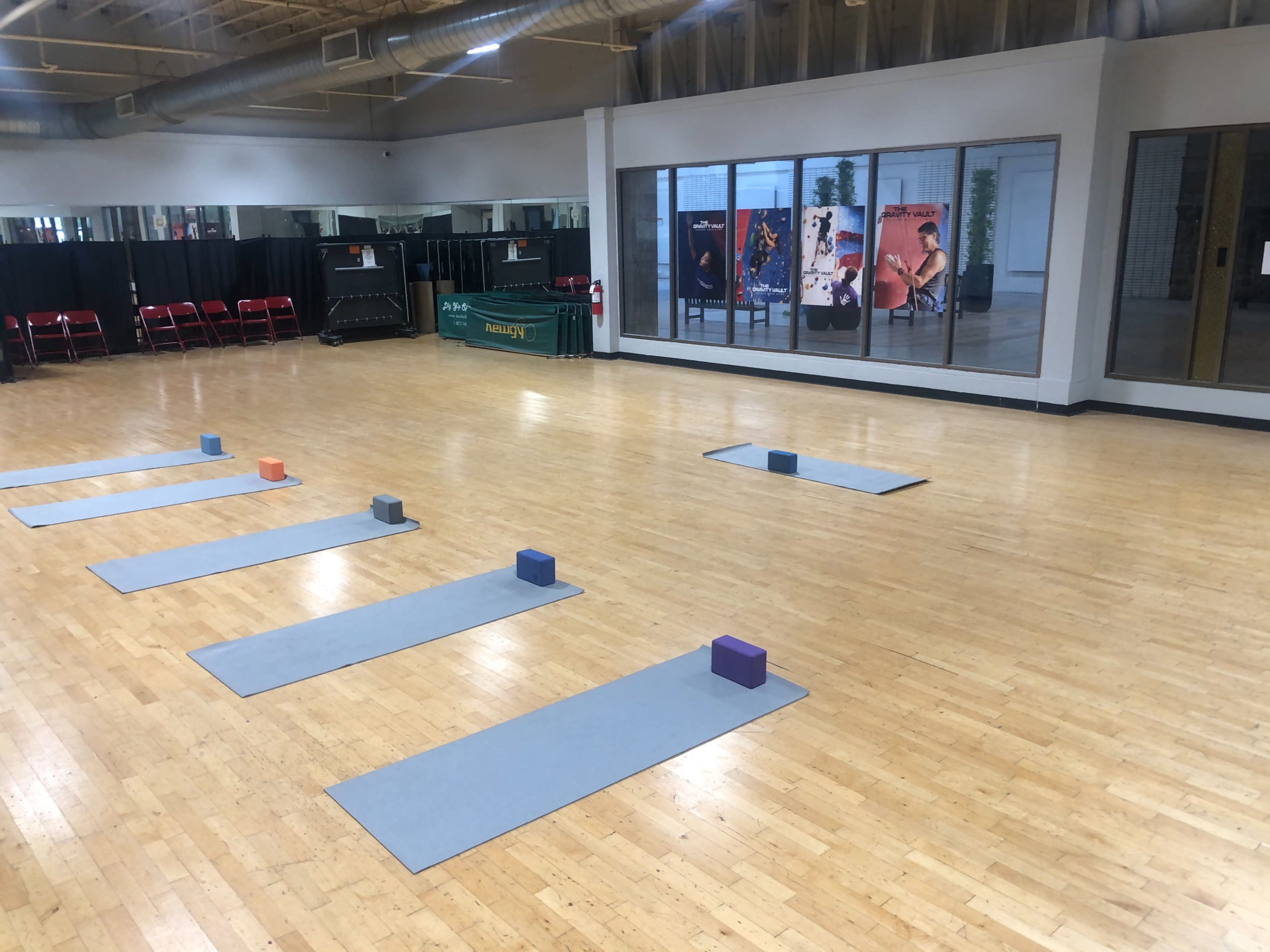 The image shows an empty studio with wooden flooring, featuring several yoga mats and blocks arranged on the floor, along with a mirrored wall and seating in the background.