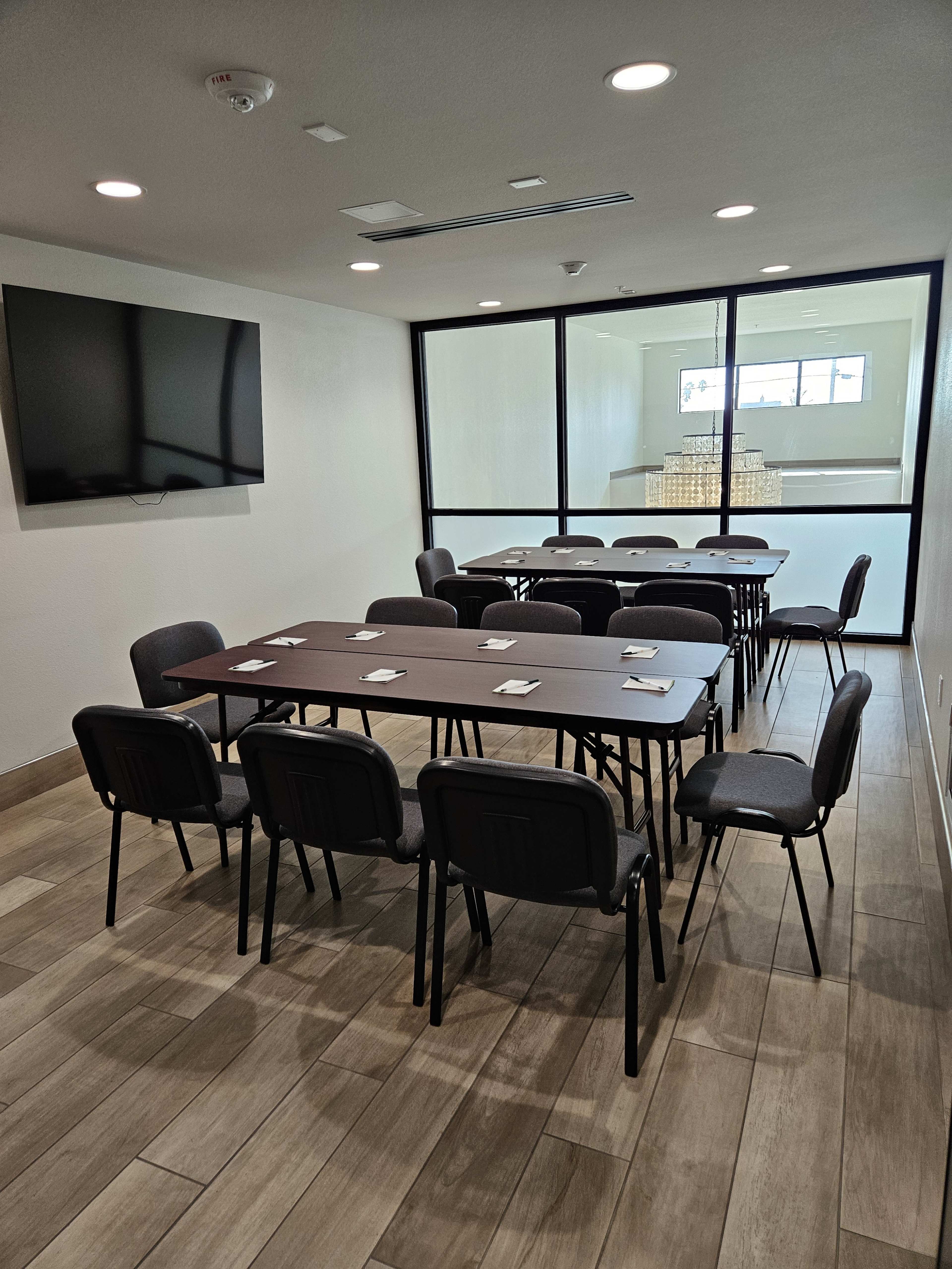 A conference room features a long table surrounded by chairs, with a television mounted on the wall and a glass partition leading to another room.