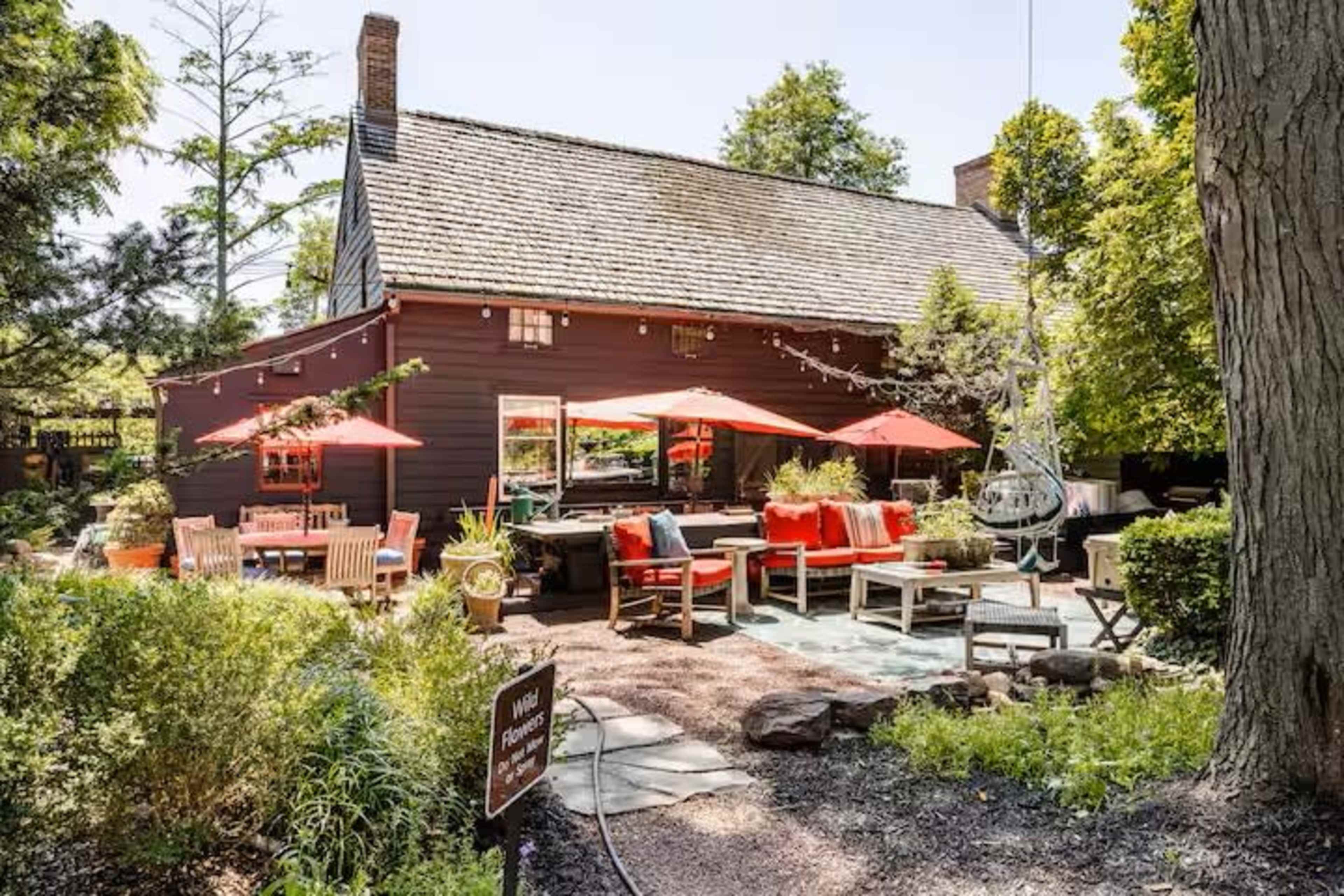 A rustic outdoor patio area features red umbrellas over seating arrangements surrounded by greenery and stone pathways at a large building.