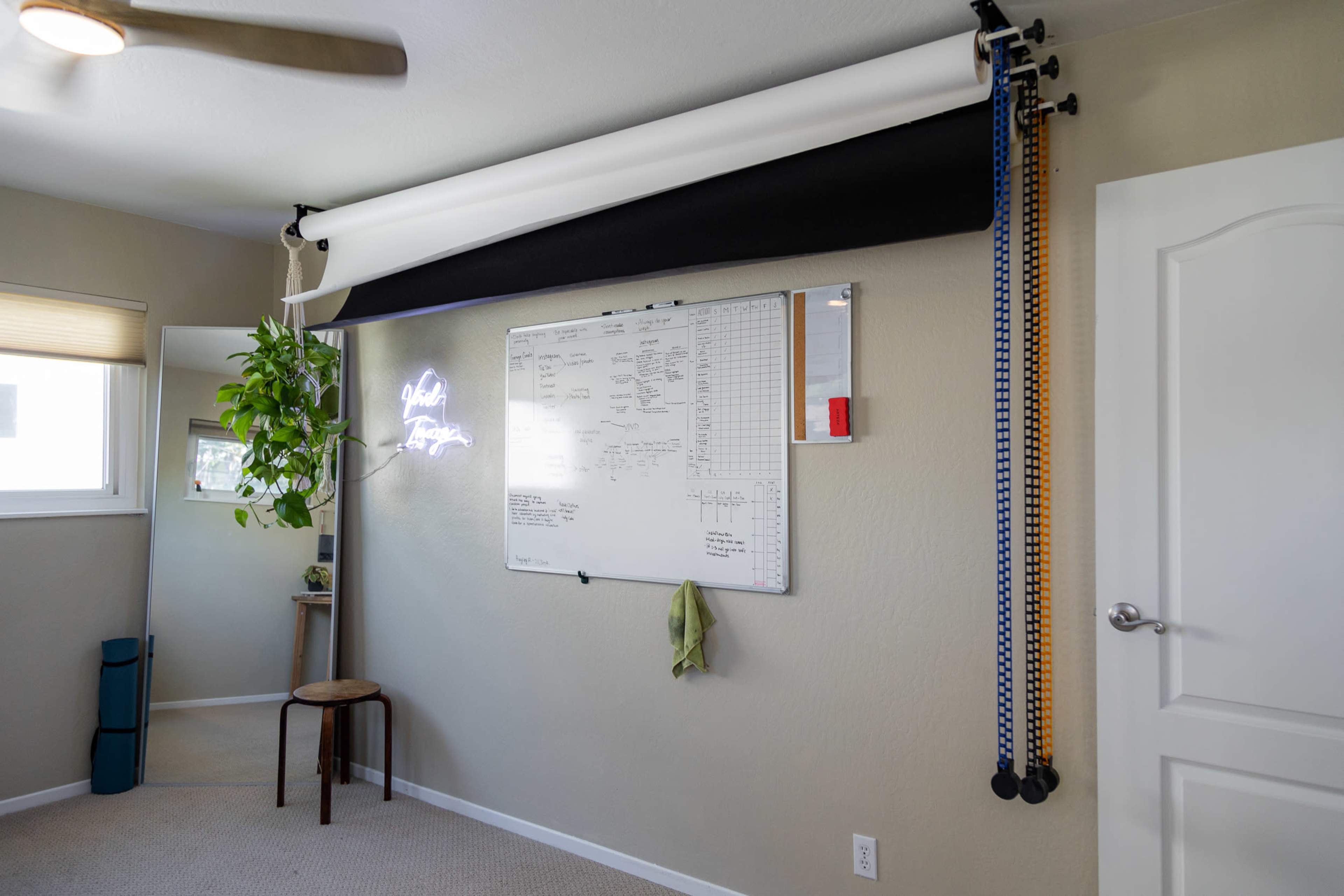A wall-mounted whiteboard with notes and a black roll-up screen is positioned next to a potted plant and a stool in a well-lit room.