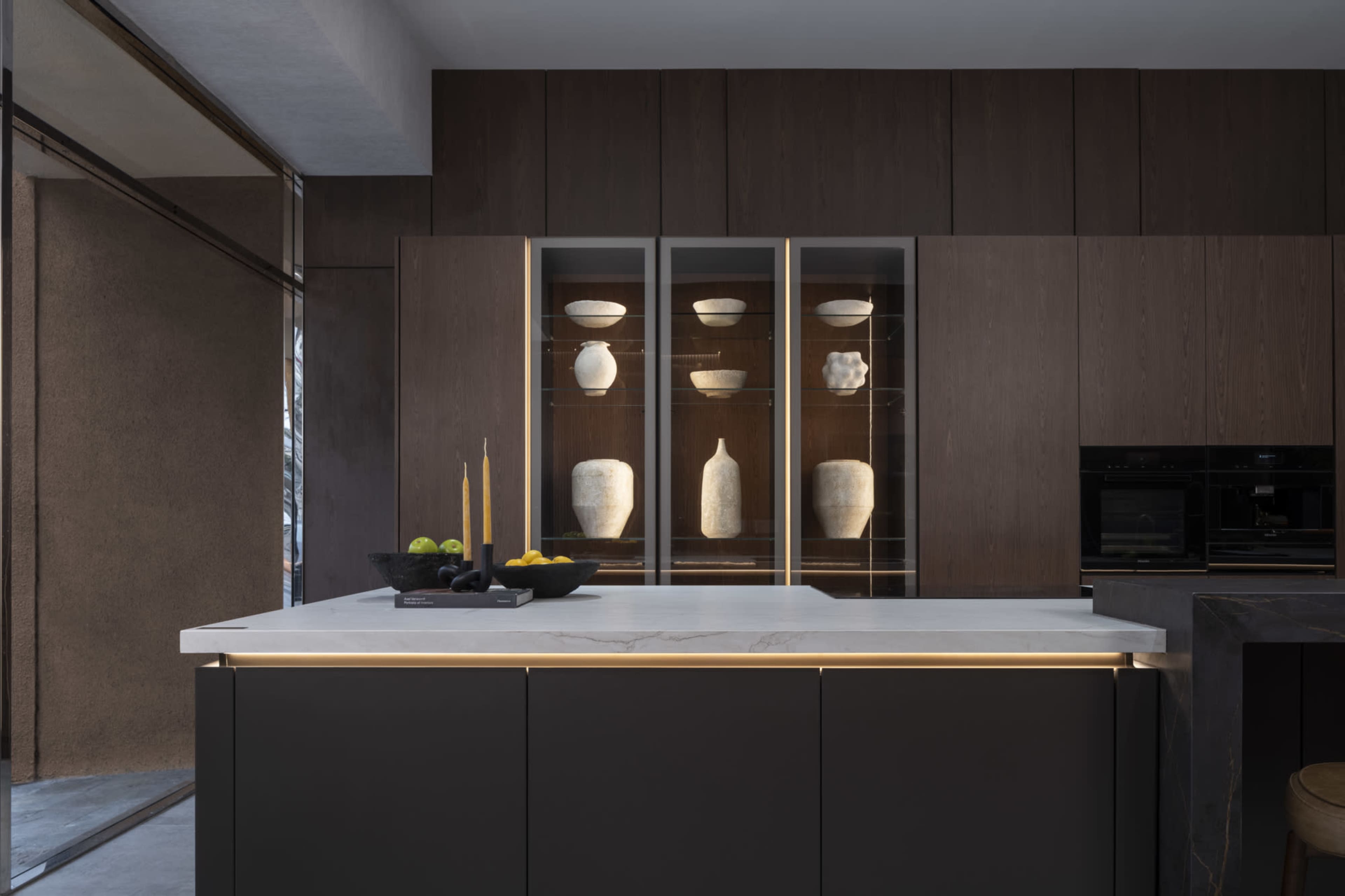 The image shows a modern kitchen featuring a minimalistic island with a white marble countertop, dark cabinetry, and glass shelves displaying decorative pottery.