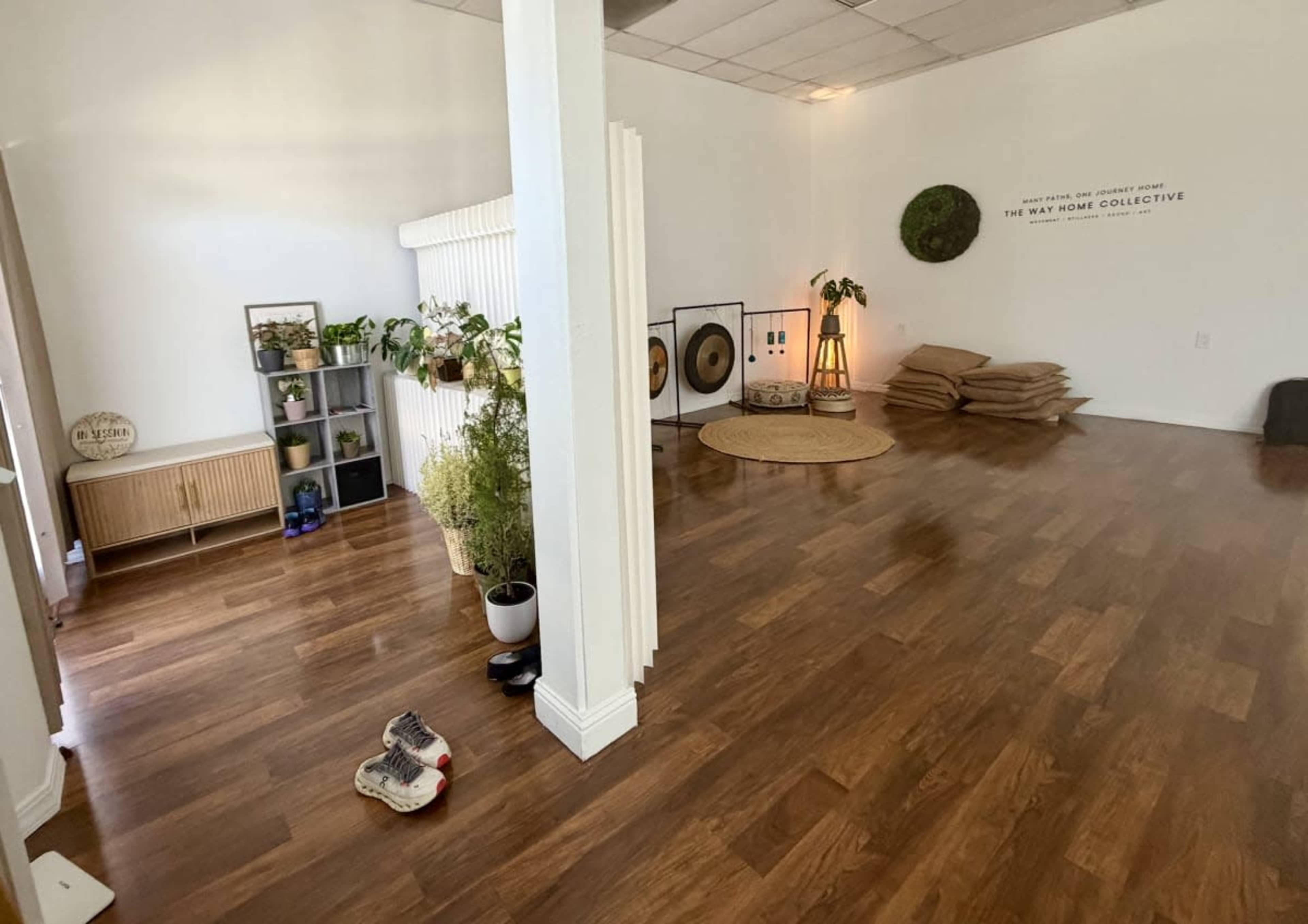 The image shows a spacious, empty room with wooden flooring, plants, and seating arrangements, along with decorative elements on the walls.