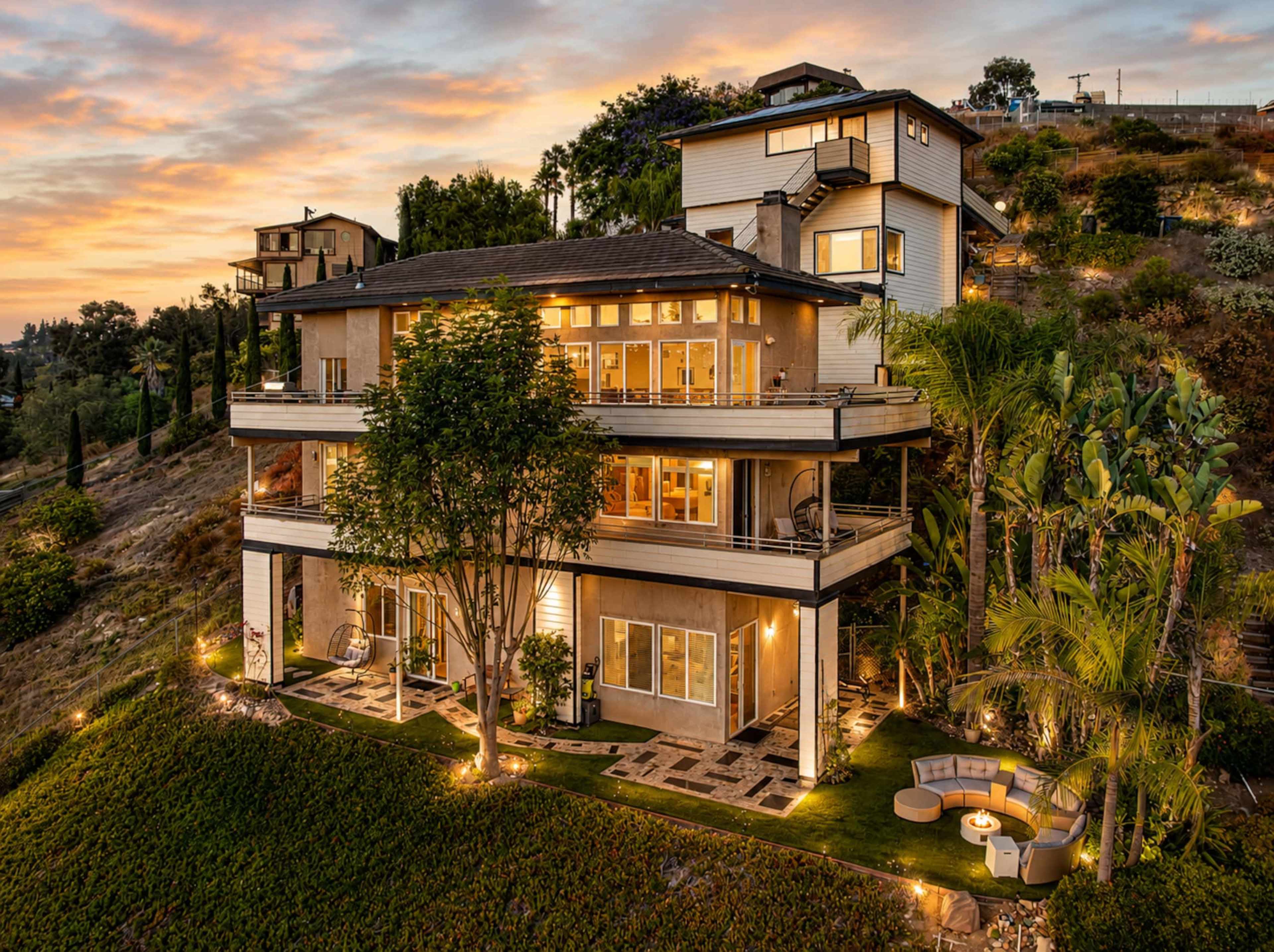 A modern three-story house is situated on a hillside, featuring large windows and a landscaped yard with seating areas, surrounded by greenery and set against a sunset sky.