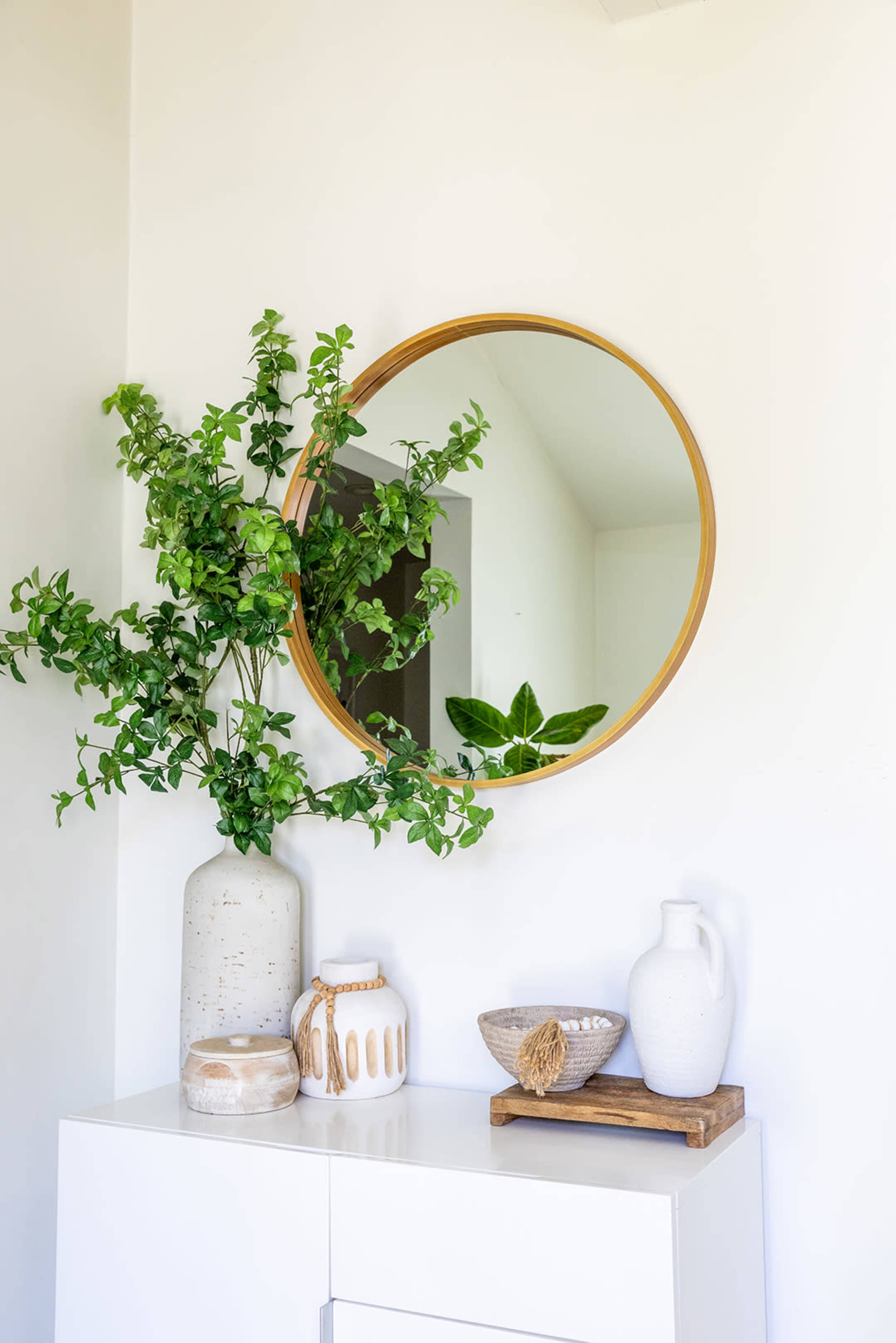 A round mirror with a wooden frame hangs above a white cabinet adorned with a vase of greenery, a decorative bowl, and a pitcher.