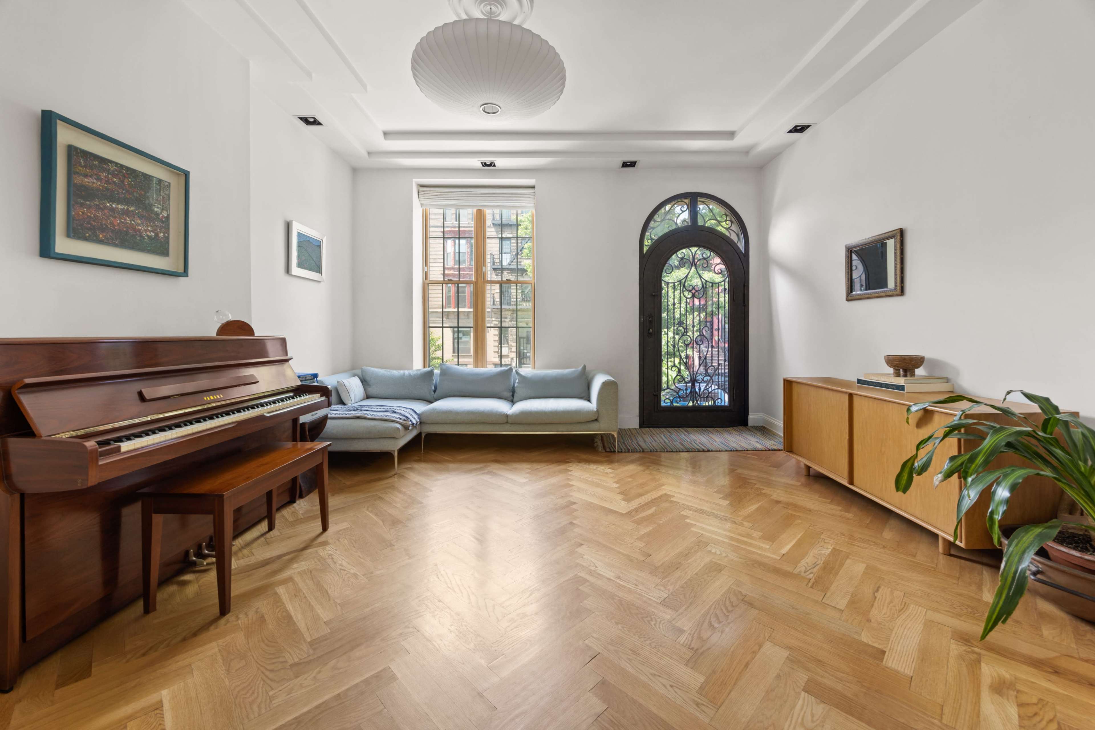 The image shows a bright living room featuring a wooden piano, a light blue sofa, a decorative wall cabinet, and a large window with a black front door in the background.