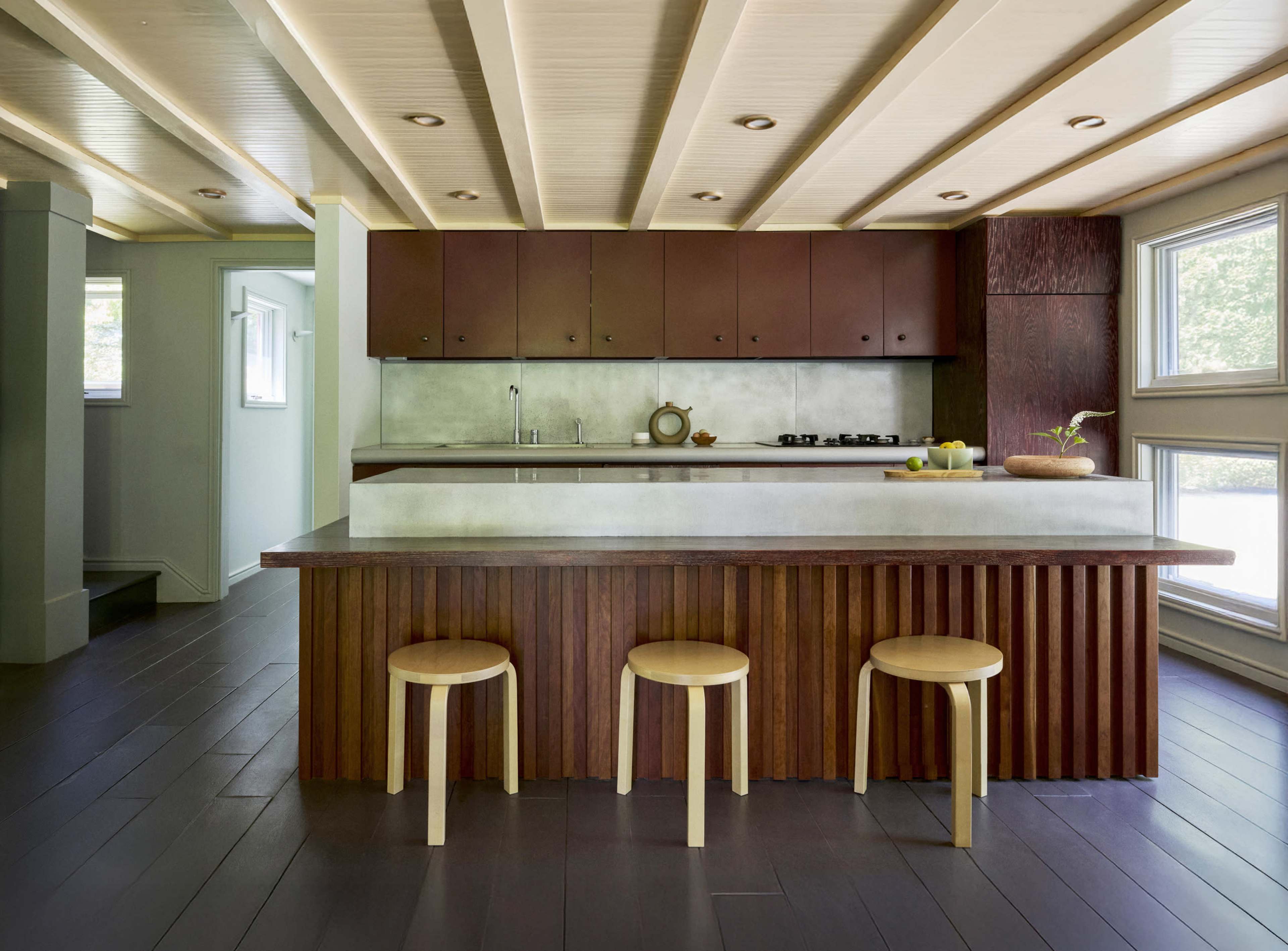 The image shows a modern kitchen with a concrete countertop, brown cabinetry, and wooden stools arranged around a central island.