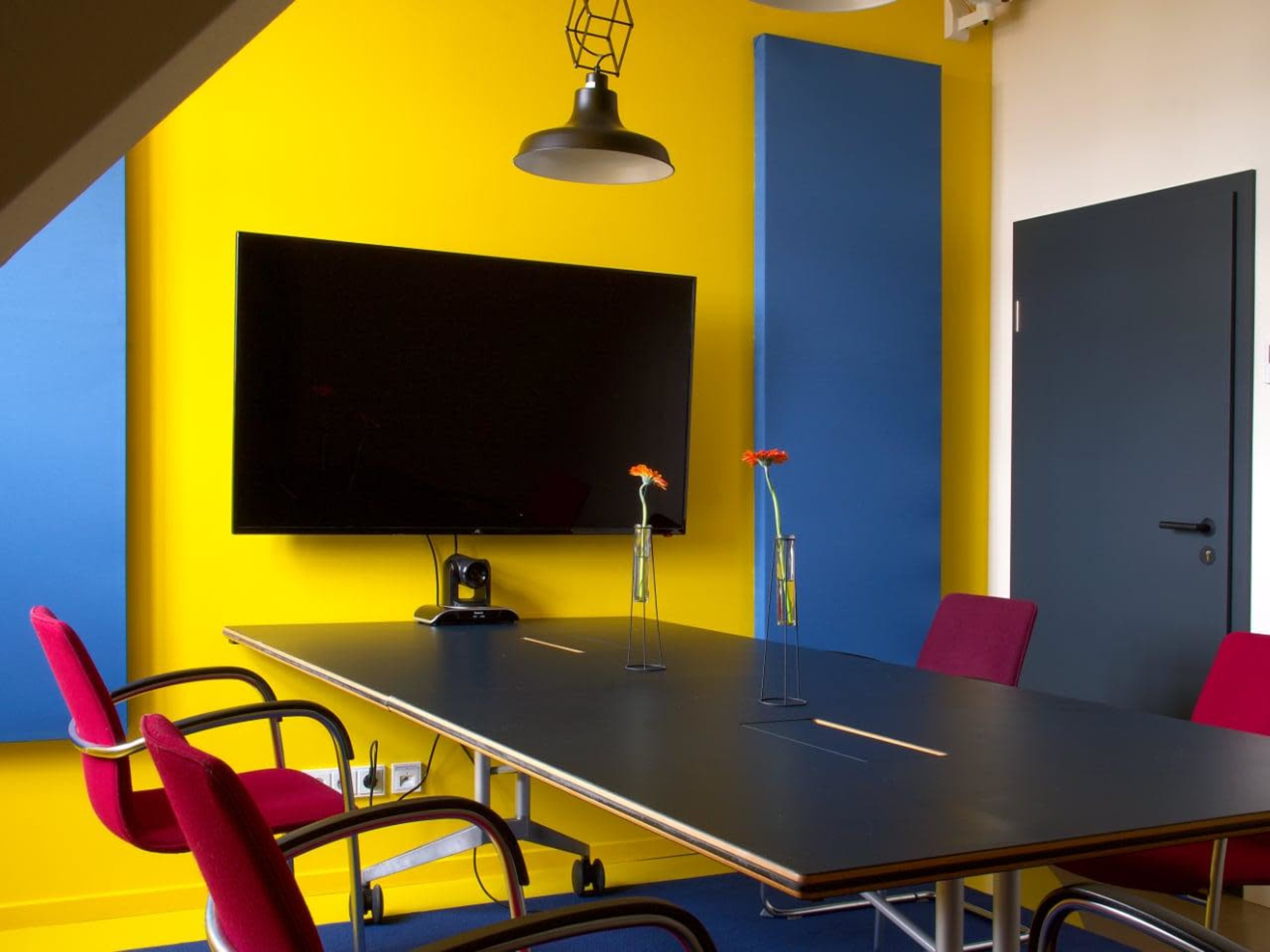 A modern conference room features a rectangular black table, colorful wall panels, a large television screen, and two pink chairs arranged around the table with small vases containing flowers.
