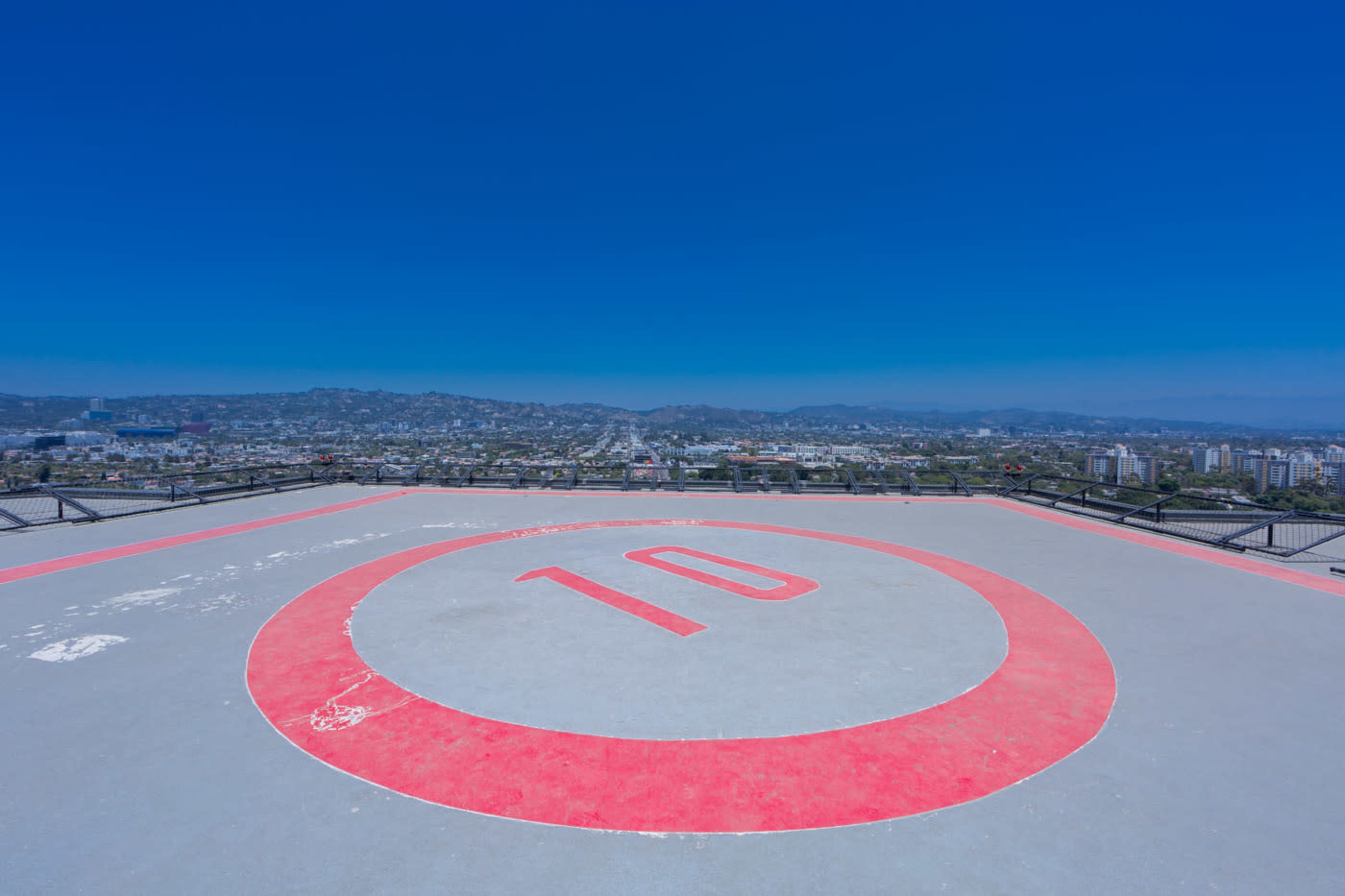 A helicopter landing pad with the number "10" painted in red is situated on a rooftop, overlooking a cityscape under a clear blue sky.