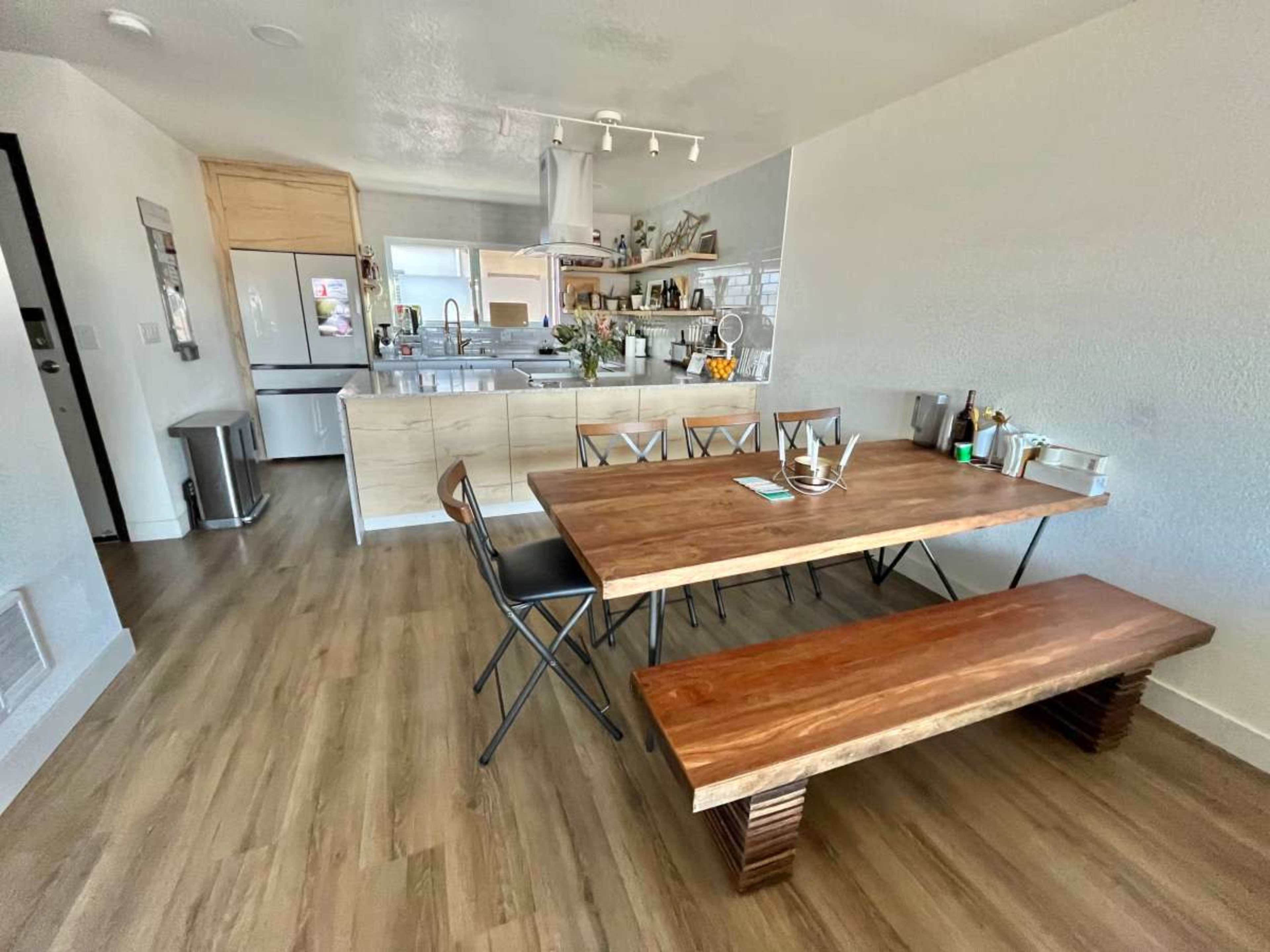 A modern dining area features a wooden table and bench, with black folding chairs, adjacent to an open kitchen with light cabinetry and appliances.