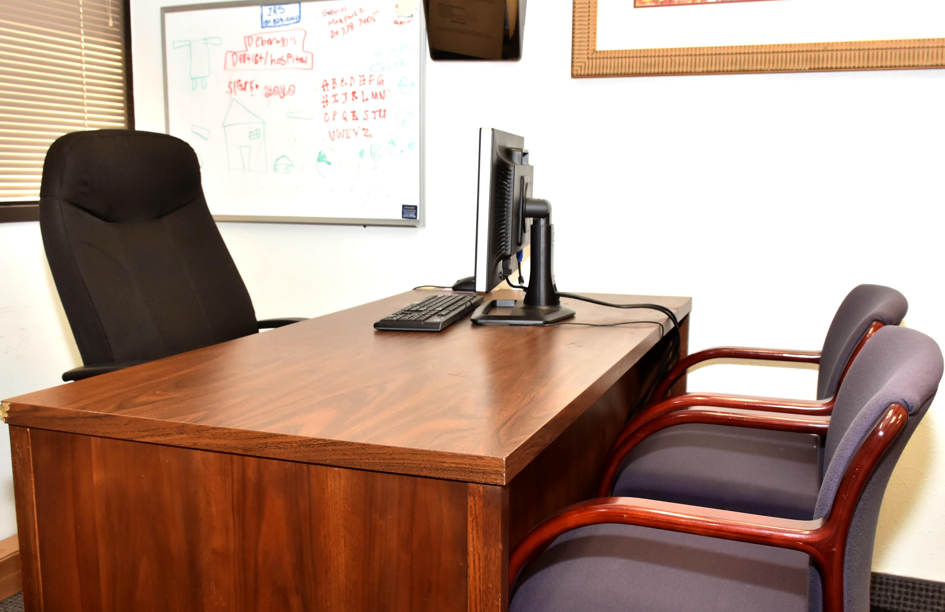 A wooden desk with a computer and keyboard, flanked by two upholstered chairs in an office setting.