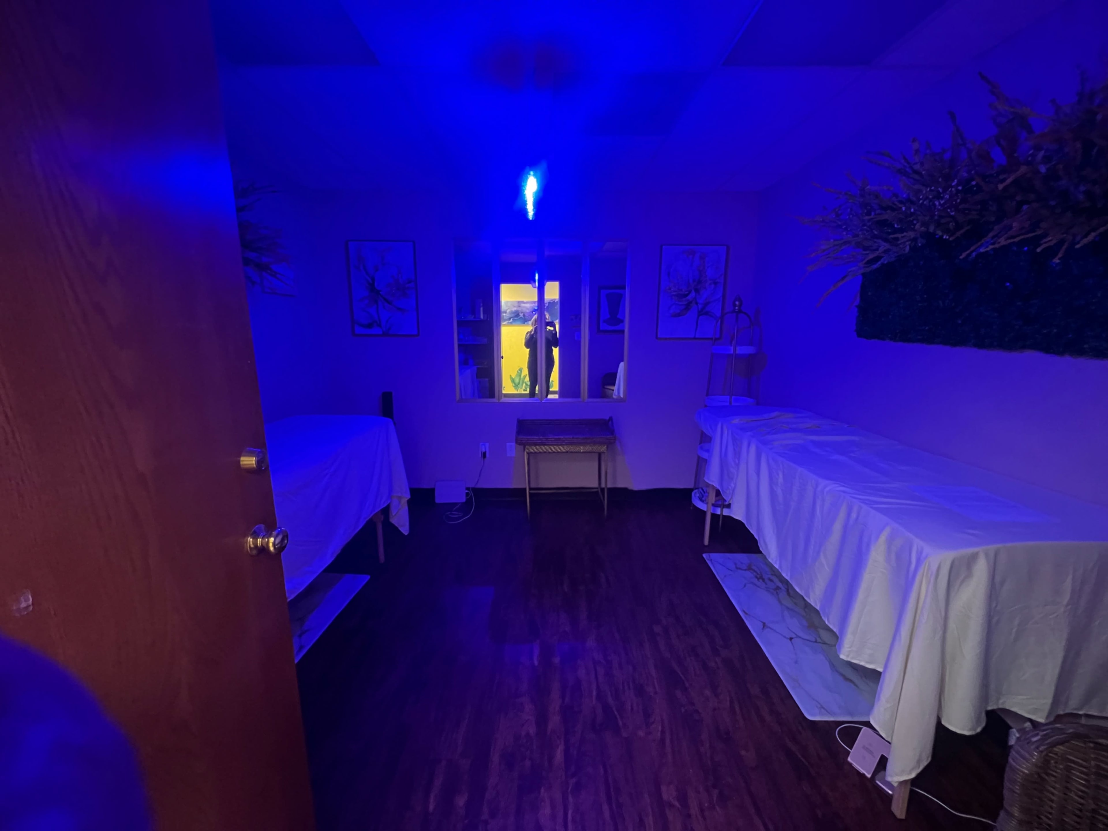 A dimly lit spa room features two massage tables covered with white linens and wall decor, with a window showing a blue glow from outside.