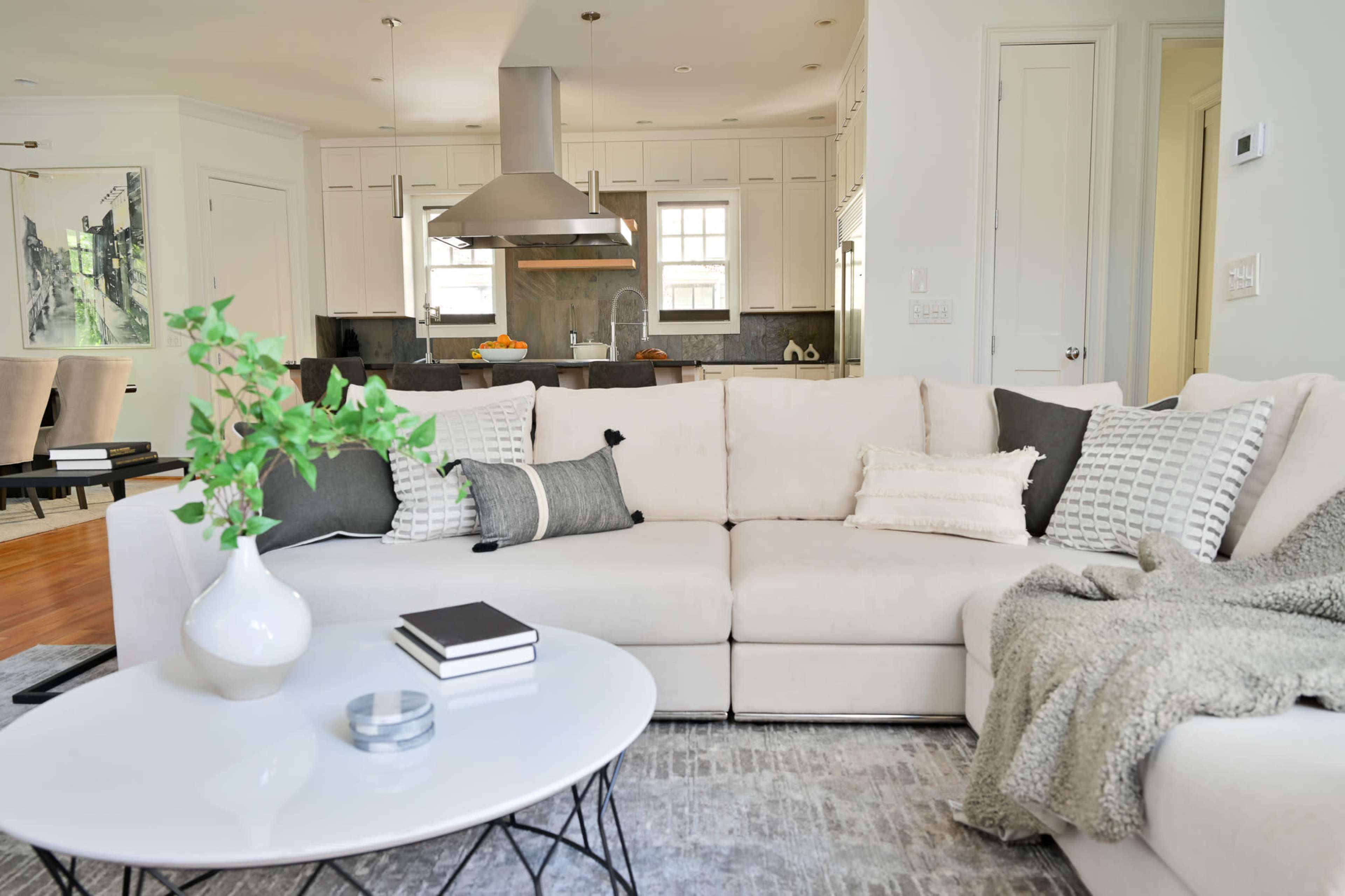 A modern living room features a light-colored sectional sofa, a round coffee table with books, and a view of a kitchen in the background.