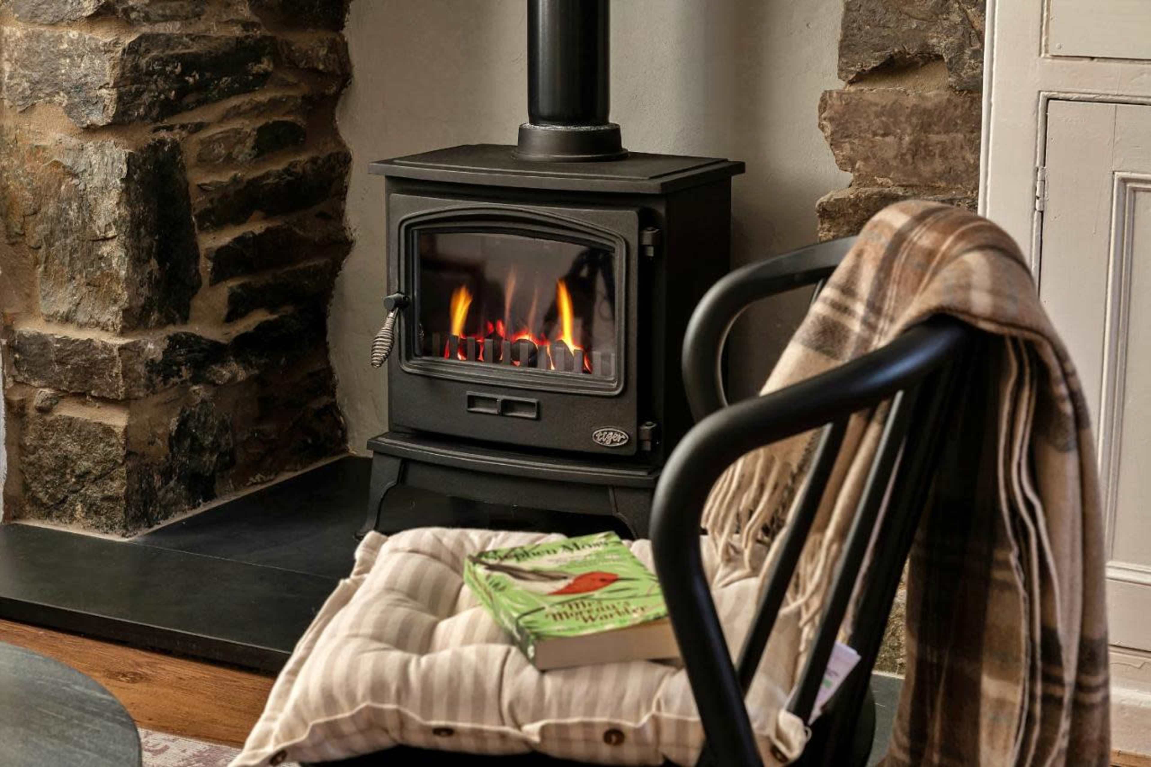 A black stove with a flame is positioned beside a stone wall, next to a chair draped with a plaid blanket and a book on its seat.