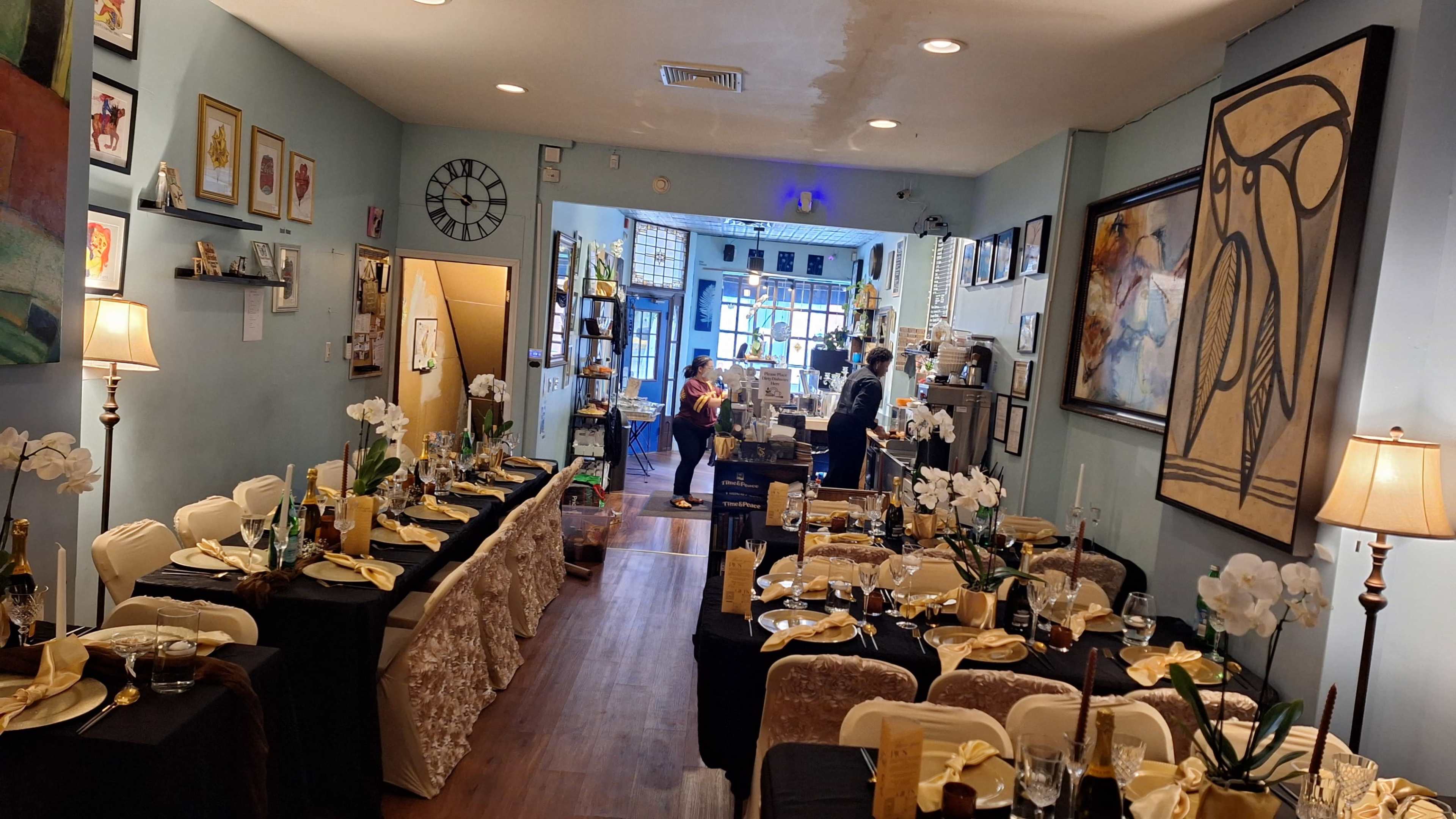 The image shows a dining area set with tables adorned in black and gold tablecloths, decorated with white flowers and candles, while a person is seen preparing food in the background.