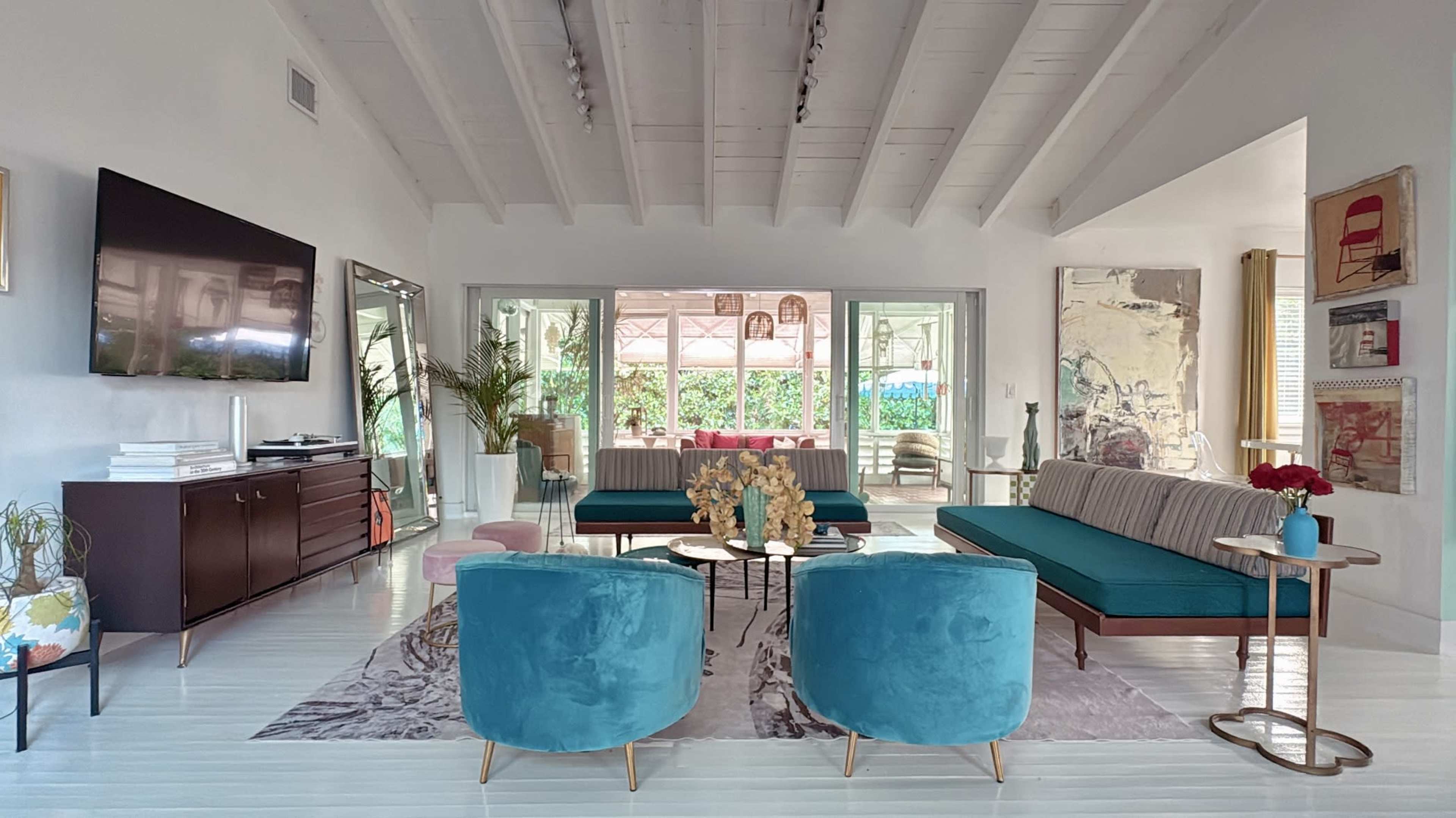 The spacious living room features a large television on the wall, two turquoise chairs, and a sleek couch, all arranged around a coffee table on a light-colored rug, with large windows providing a view of the outdoors.