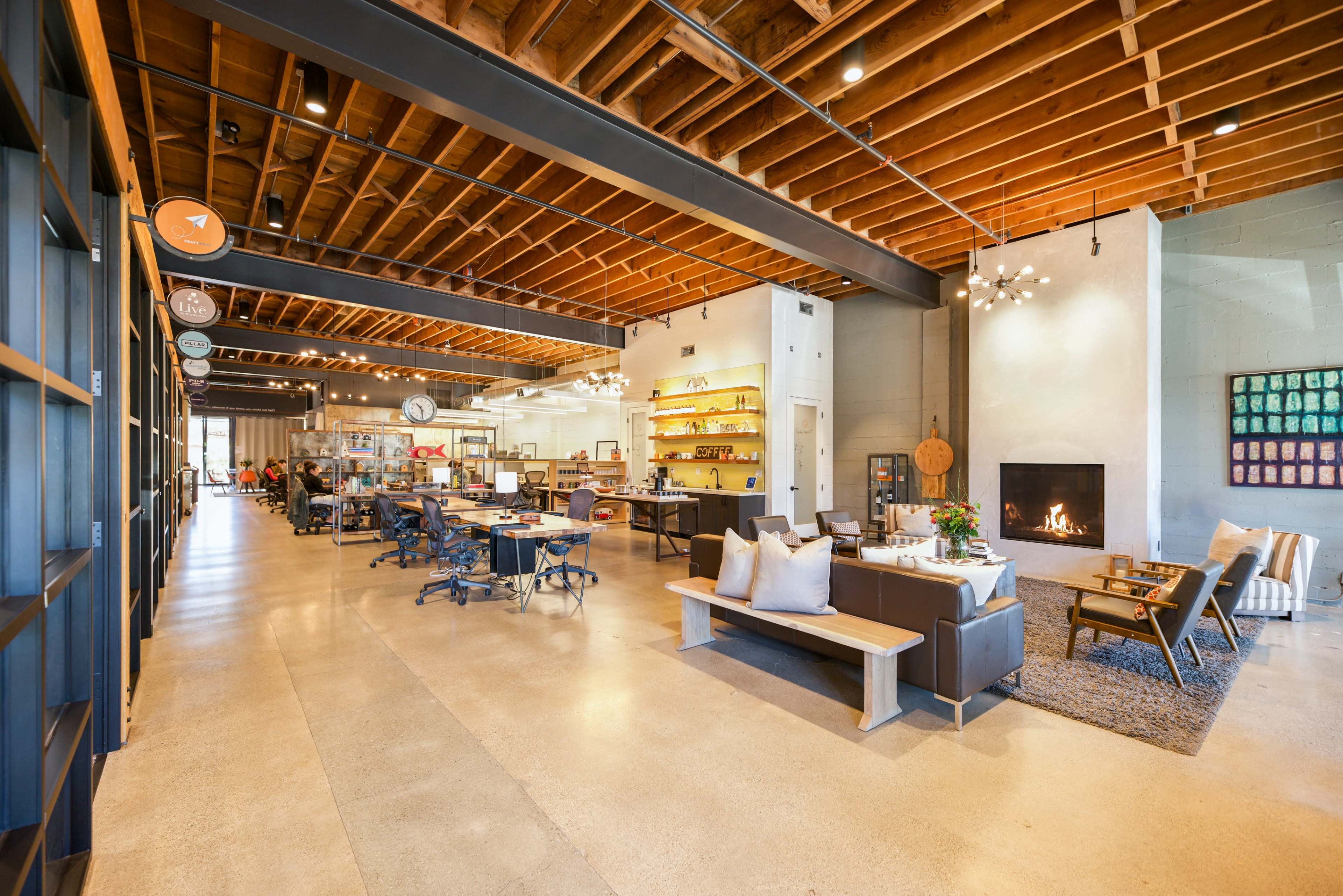 A modern, open-concept workspace featuring seating areas, a fireplace, and a kitchen area with industrial design elements and wooden beams overhead.
