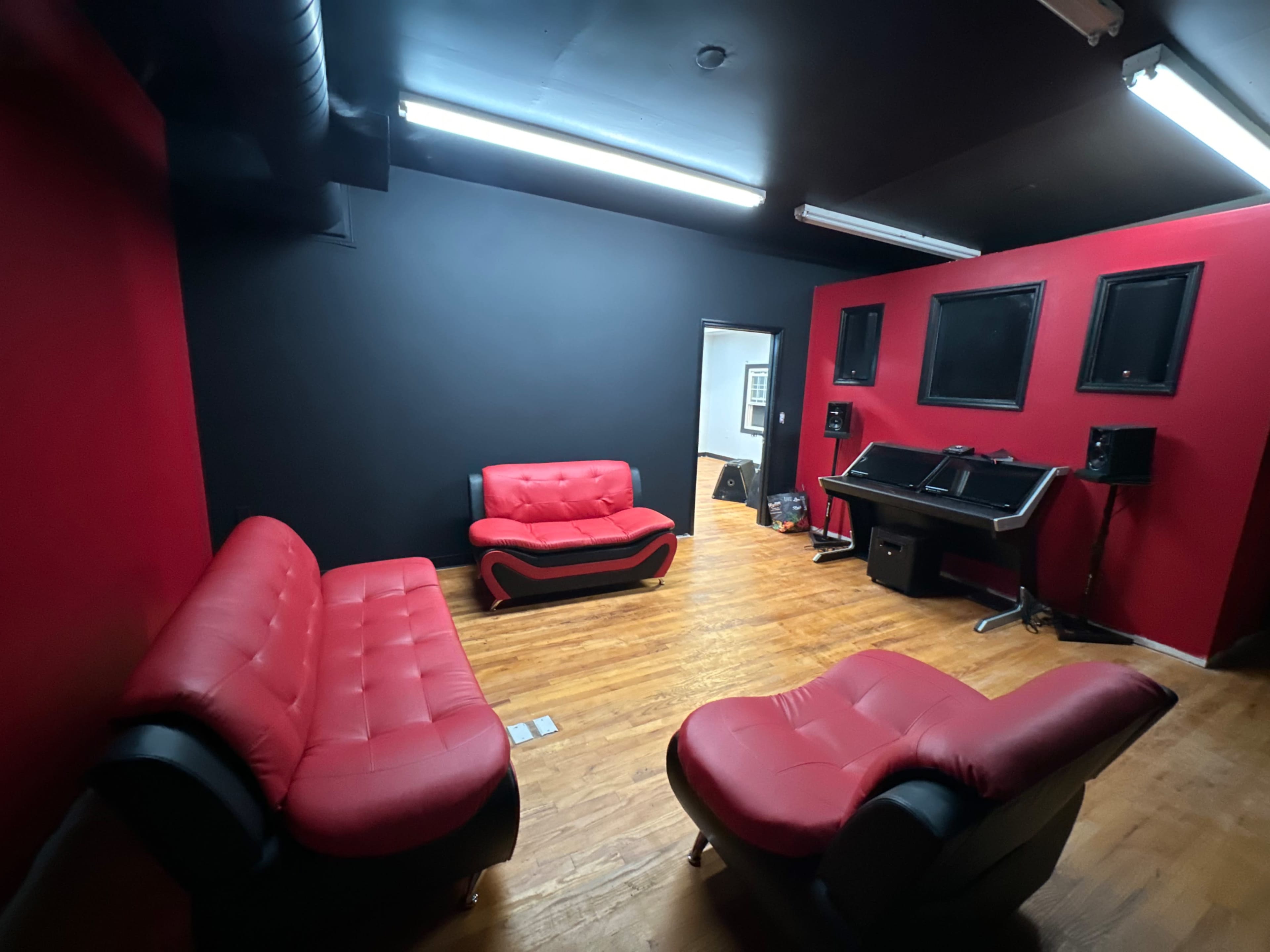 The image shows a modern room with red and black walls, featuring black leather furniture, a desk with audio equipment, and wooden flooring.