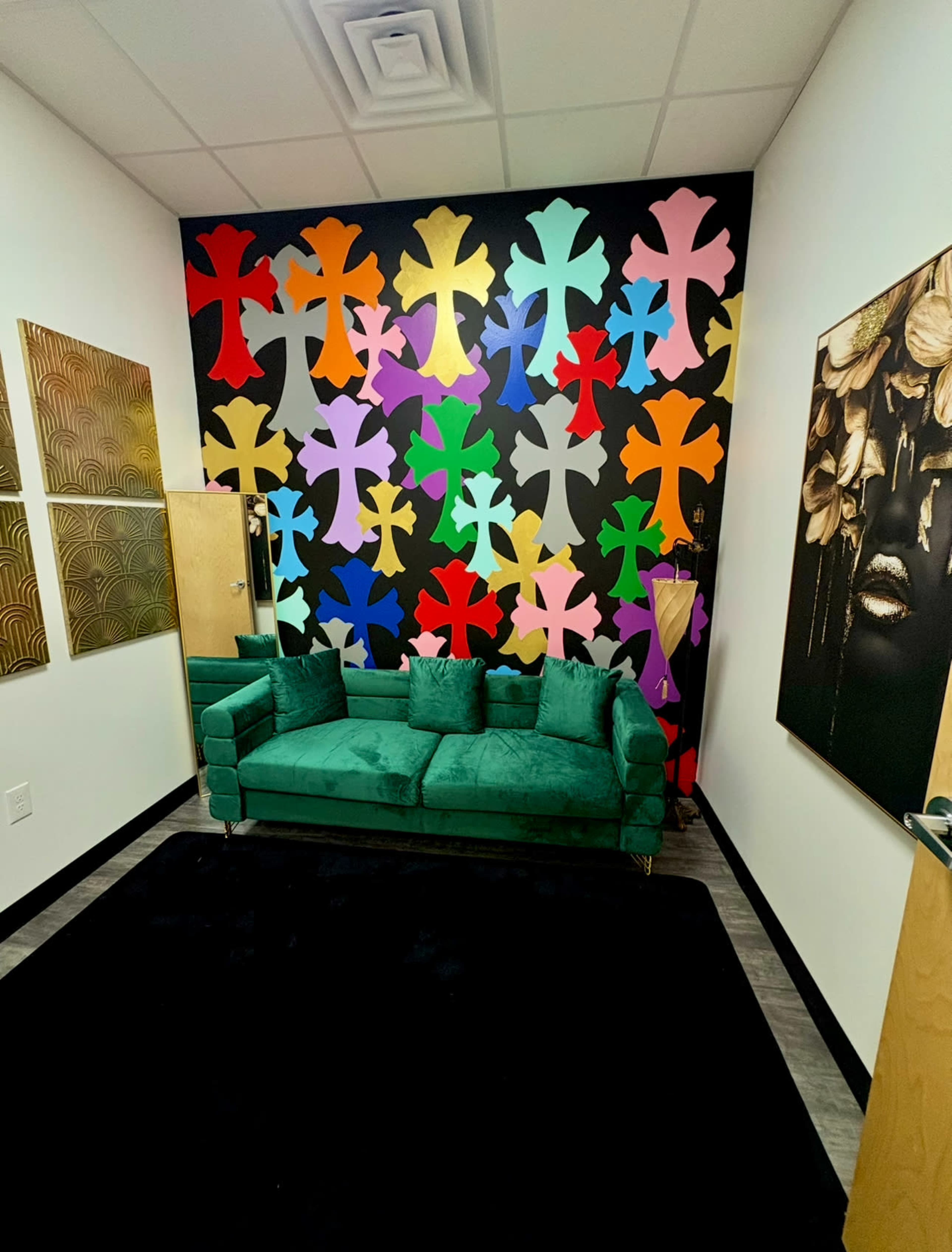 The image shows a small room featuring a green velvet sofa, a black rug, and a brightly colored wall adorned with various cross shapes in multiple colors.
