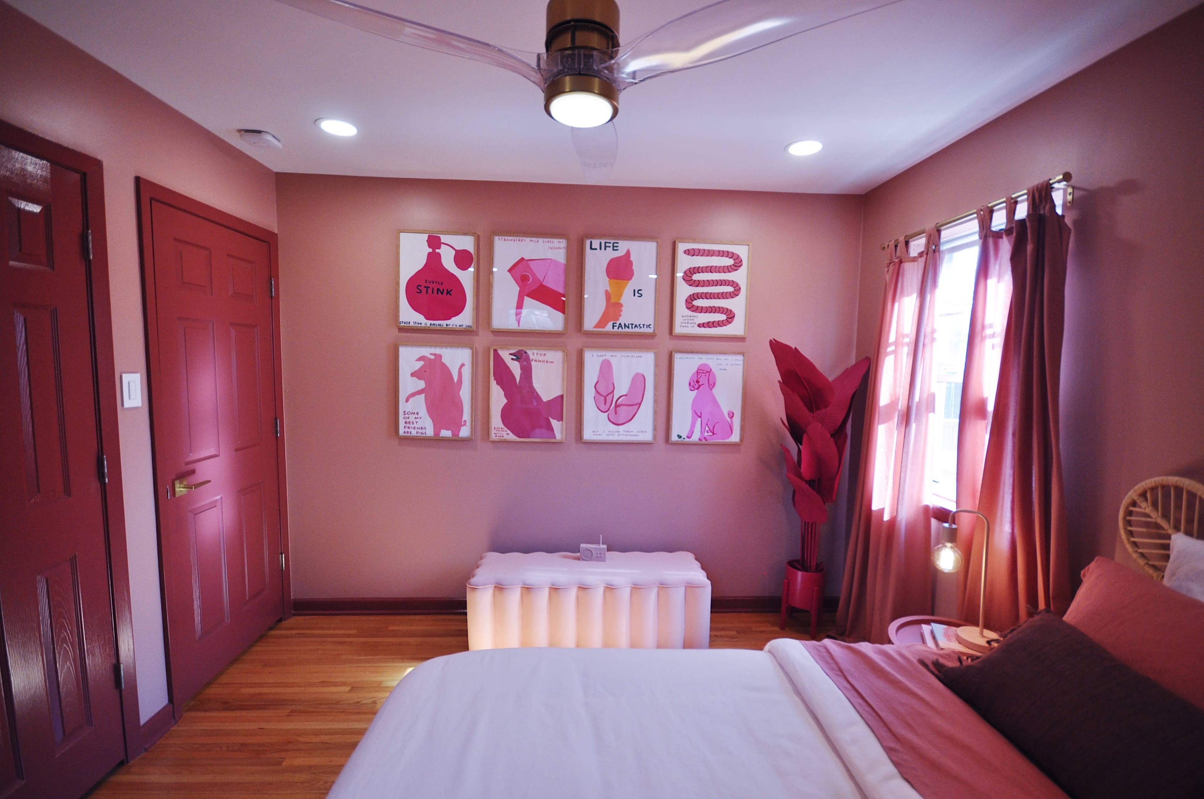 A bedroom features pink walls, a bed with white bedding, a modern light fixture, and framed art pieces on the wall.