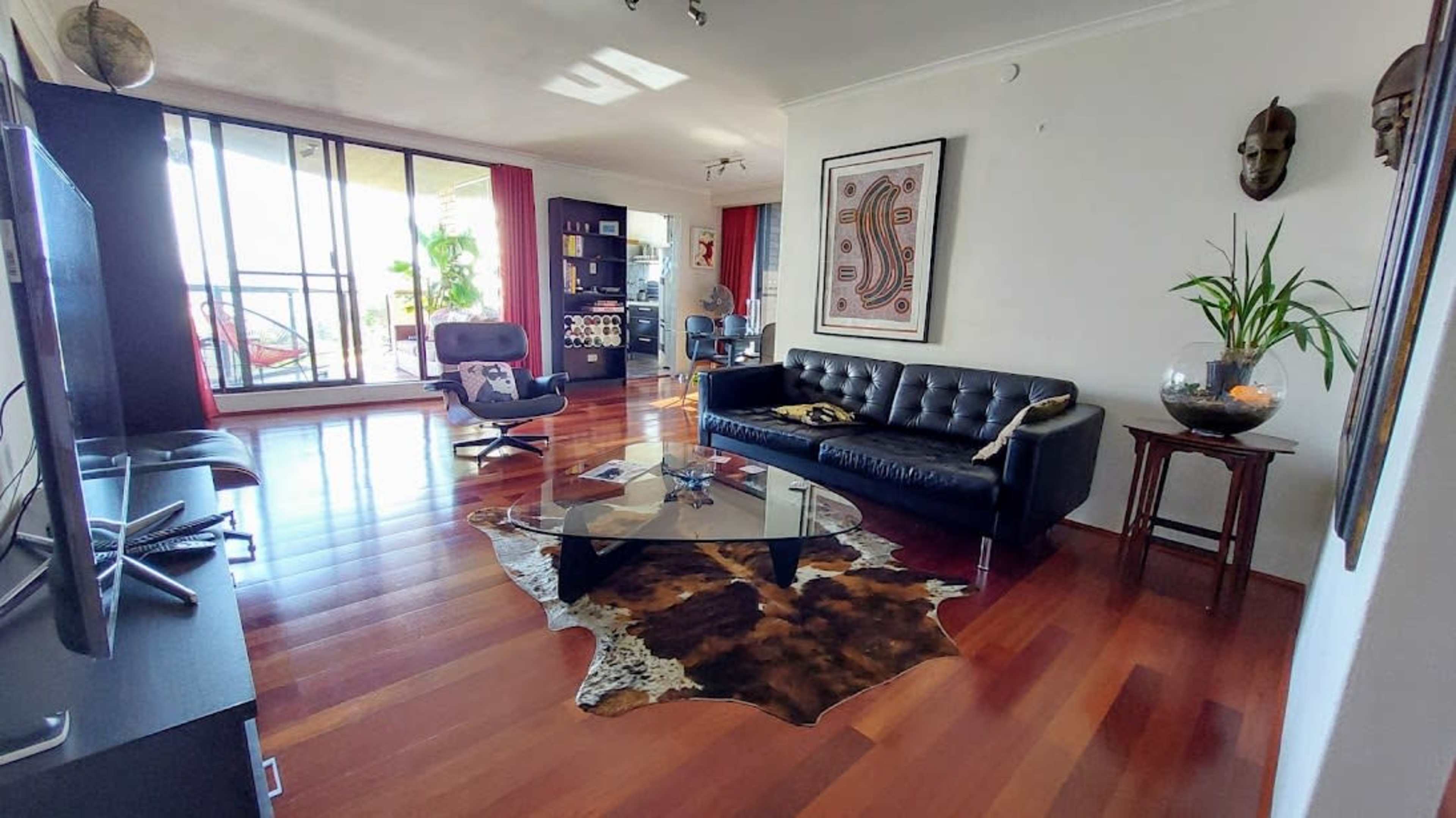 A modern living room featuring a black leather sofa, a glass coffee table, wooden flooring, and large windows with red curtains.