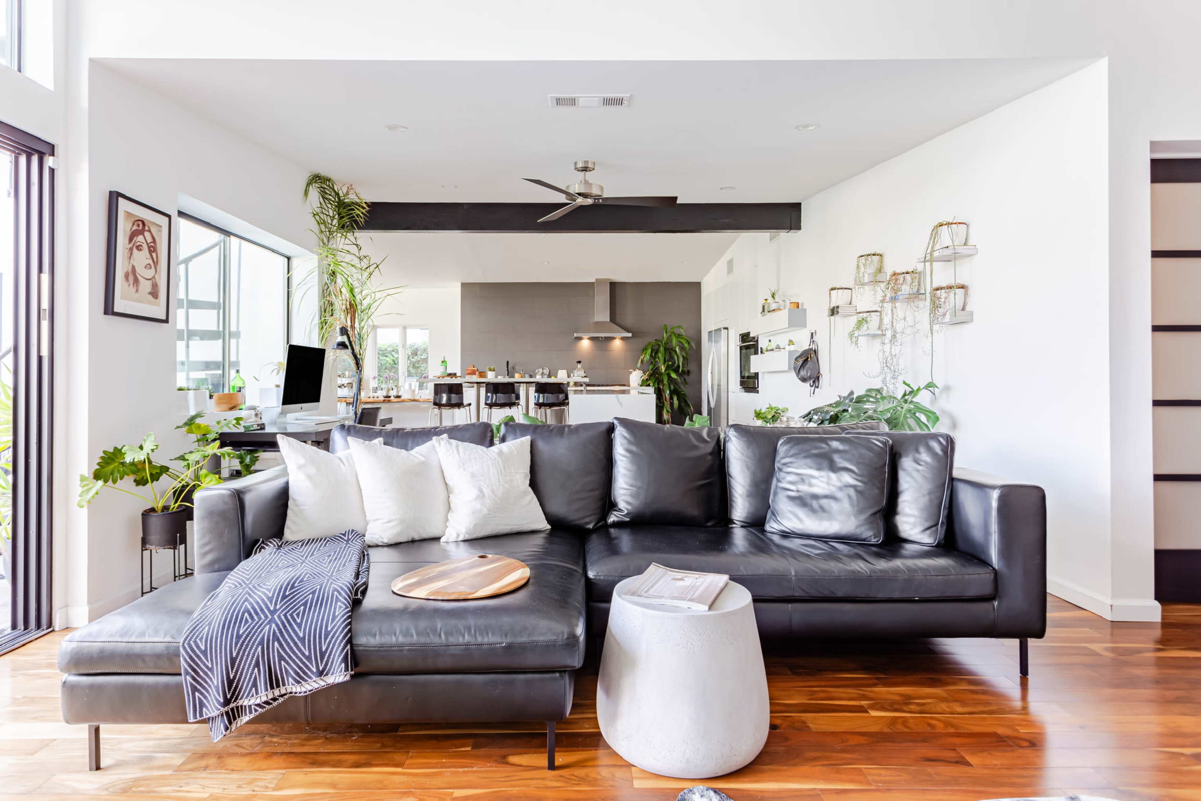 A modern living room features a black sectional sofa with decorative pillows and a light-colored side table, overlooking an open kitchen space.