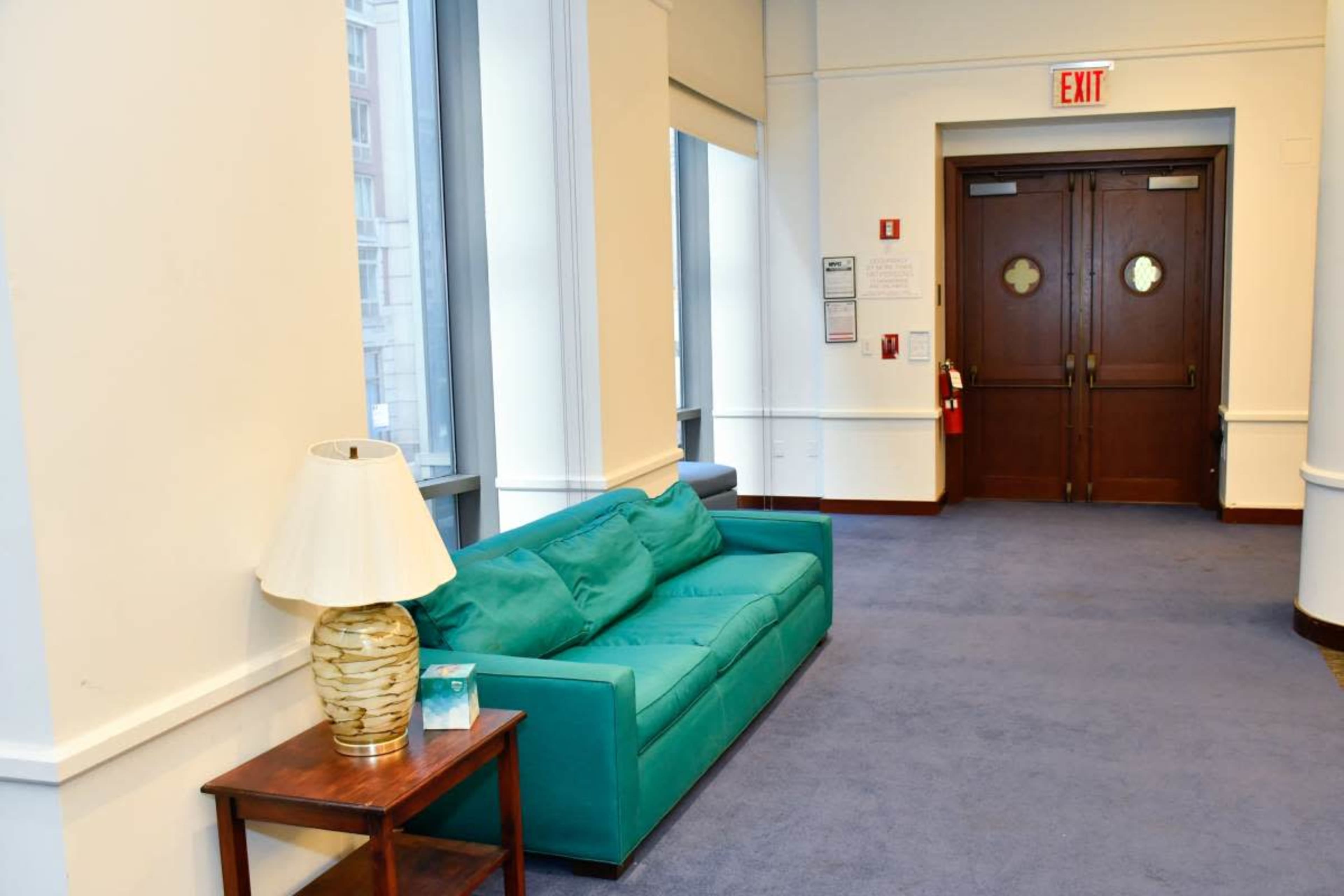 A teal couch is positioned along a wall next to a lamp and a wooden table, facing a set of double doors at the end of the hallway.