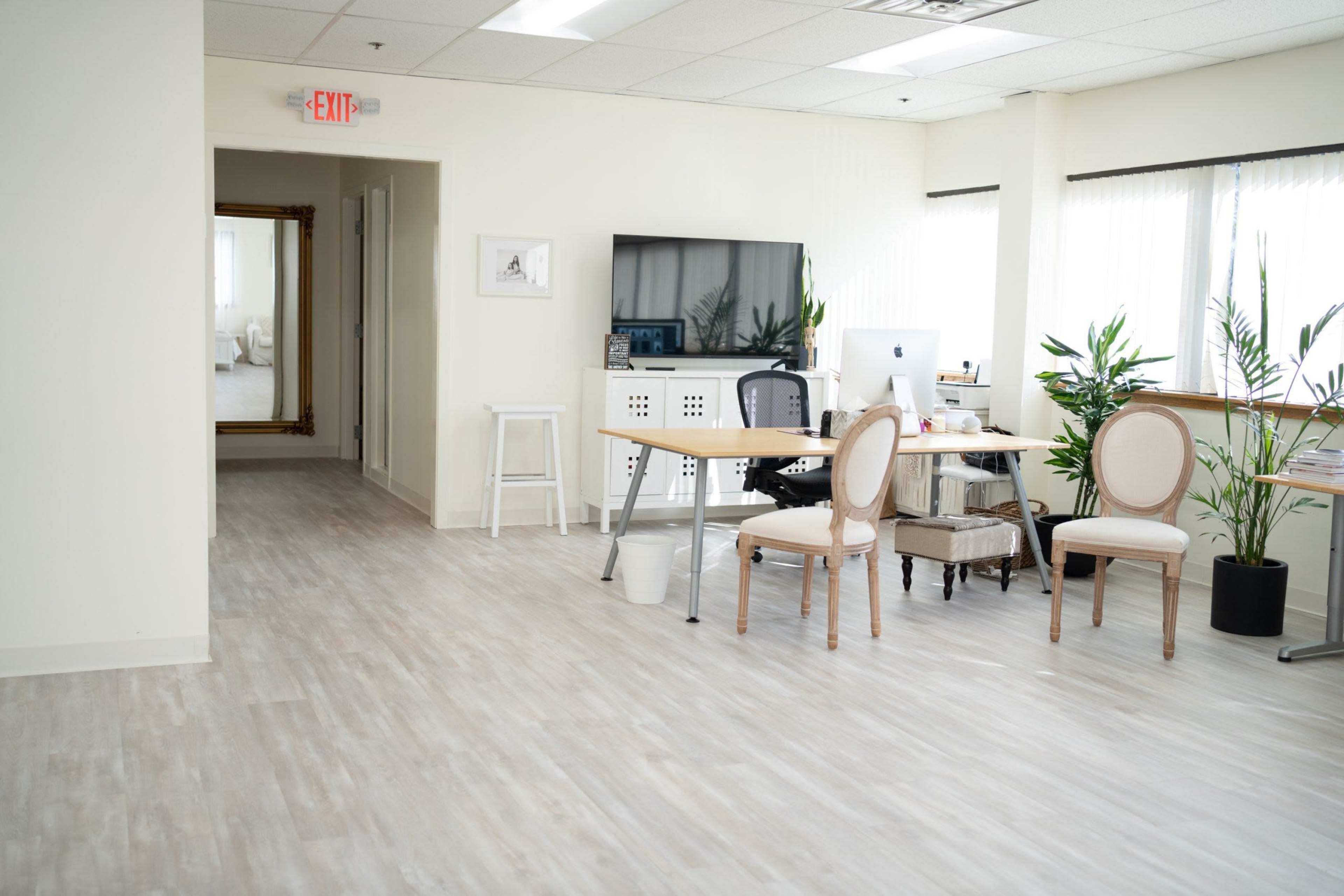 A bright office space with a desk, two chairs, a large monitor, and plants, along with a hallway leading to a mirror at the far end.