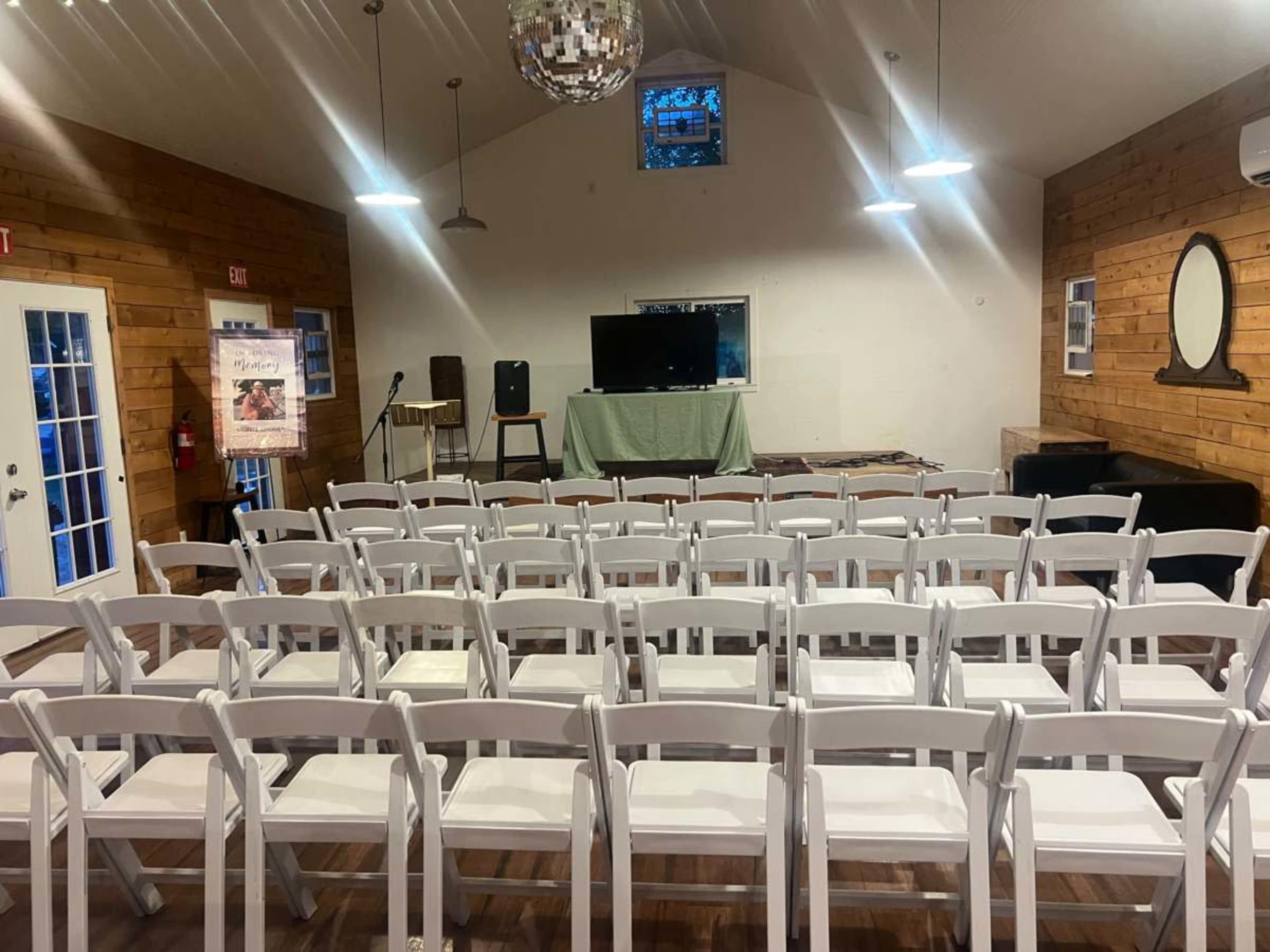 The image shows a room set up for an event with rows of white folding chairs facing a stage area that includes a TV and a small microphone stand.