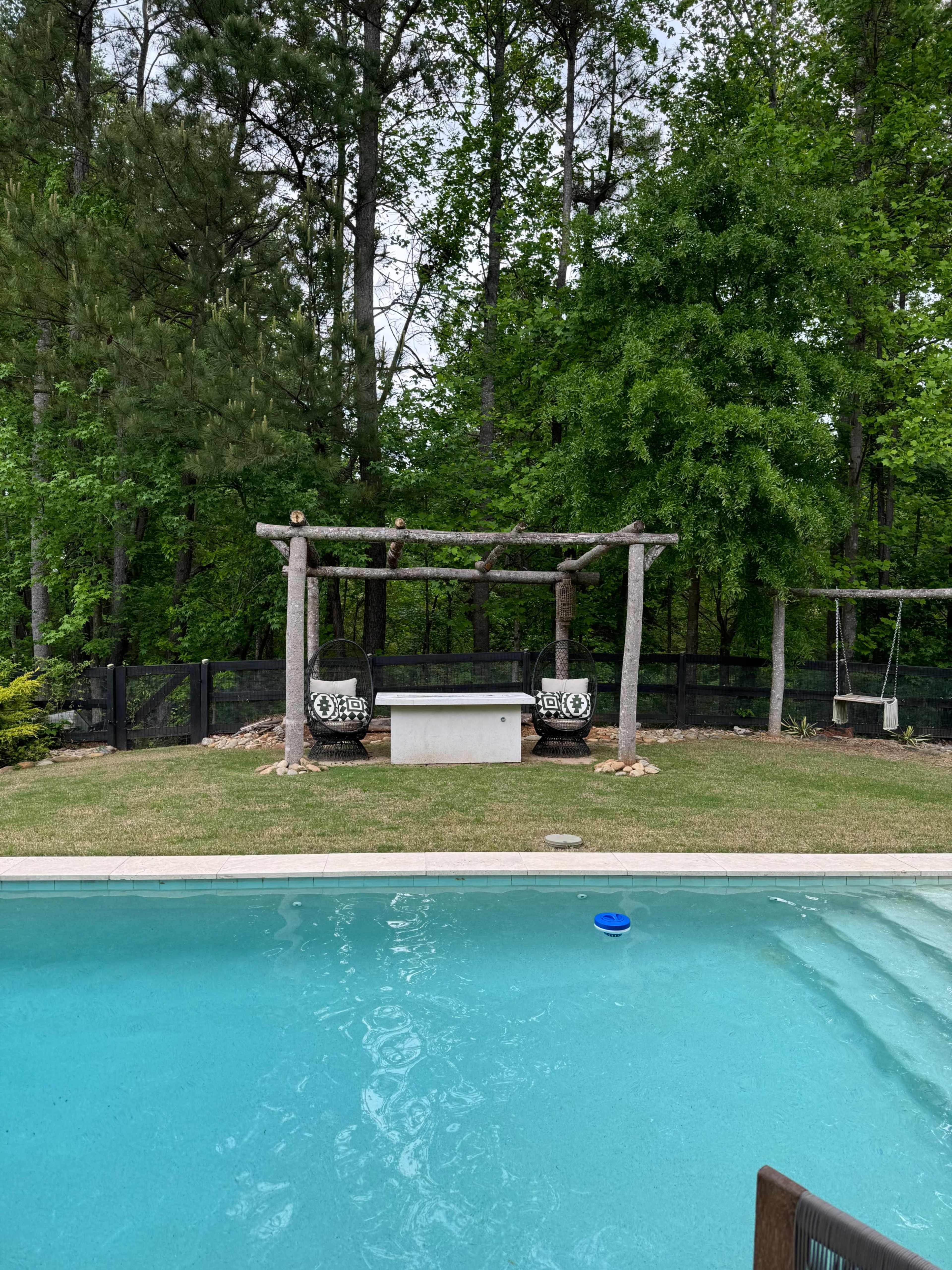 The image shows a swimming pool with clear blue water, framed by a wooden pergola and surrounded by green trees.