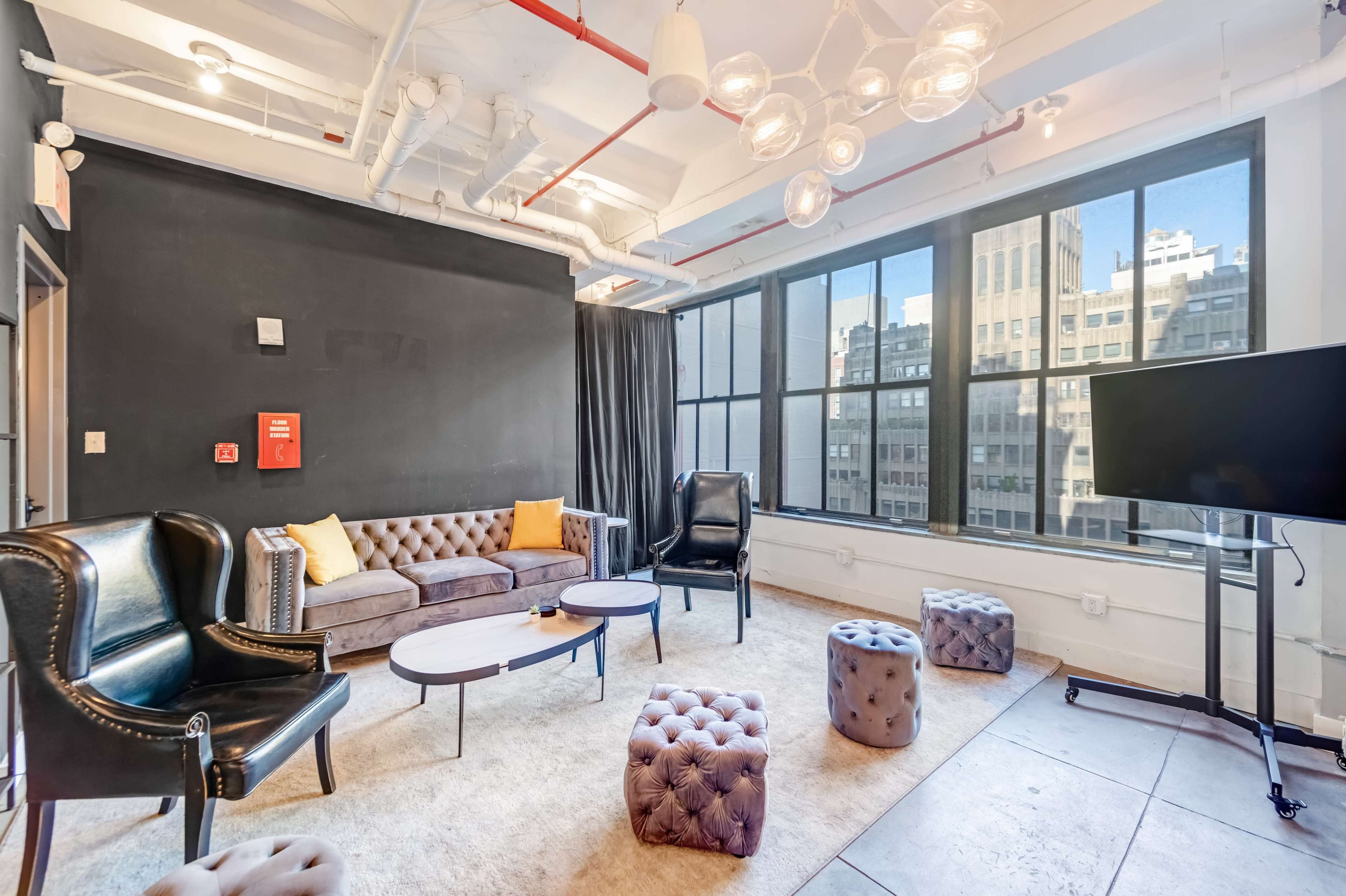 The room features a seating area with a brown leather sofa, armchairs, and tufted ottomans, surrounded by large windows and modern lighting fixtures.