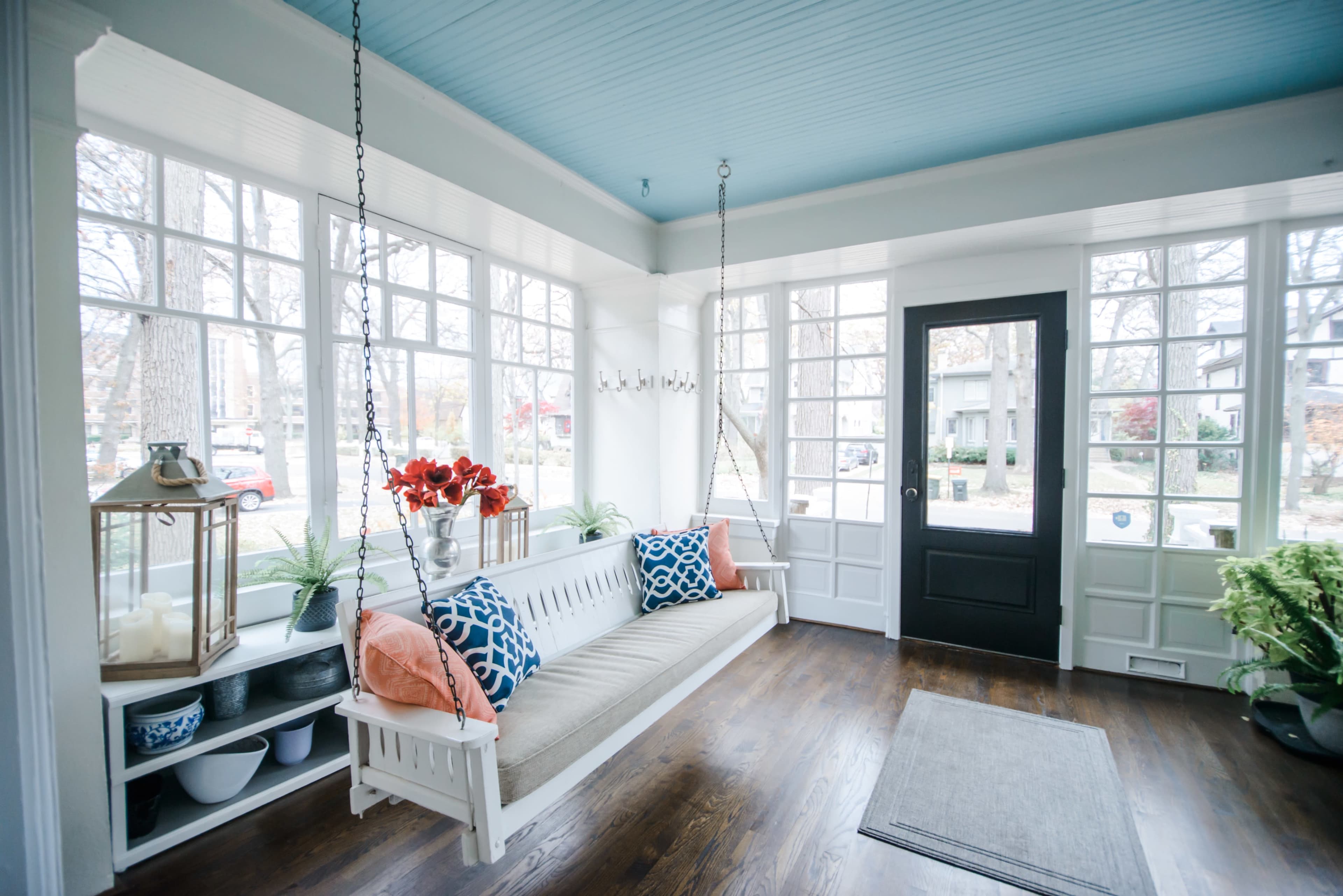 A sunlit porch featuring a white swing bench, blue ceiling, and large windows adorned with plants and decorative pillows.