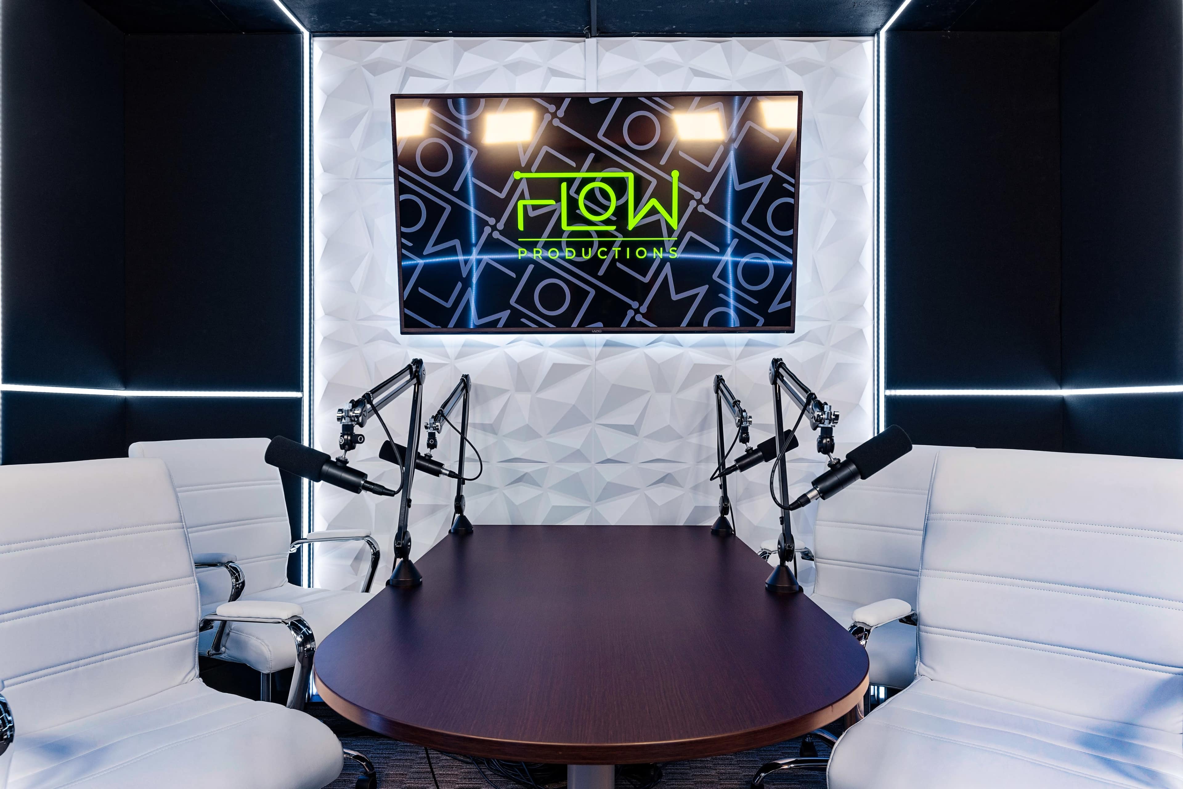 A modern recording studio with a round table, white chairs, and microphones set up, featuring a large screen displaying the logo for "Flow Productions" on the wall.