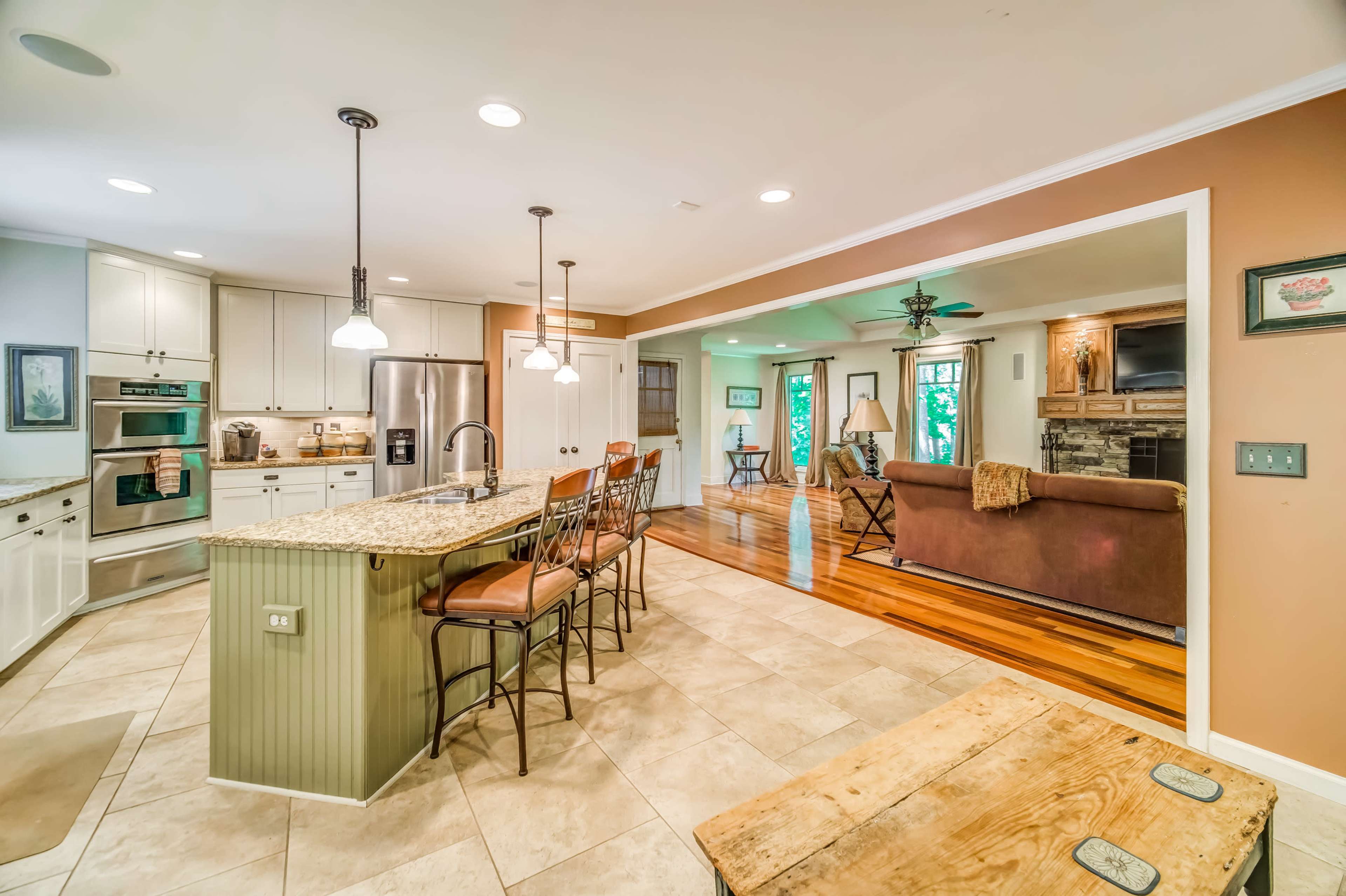 The image shows an open kitchen and living area with a granite countertop island, tiled flooring, and wooden accents.