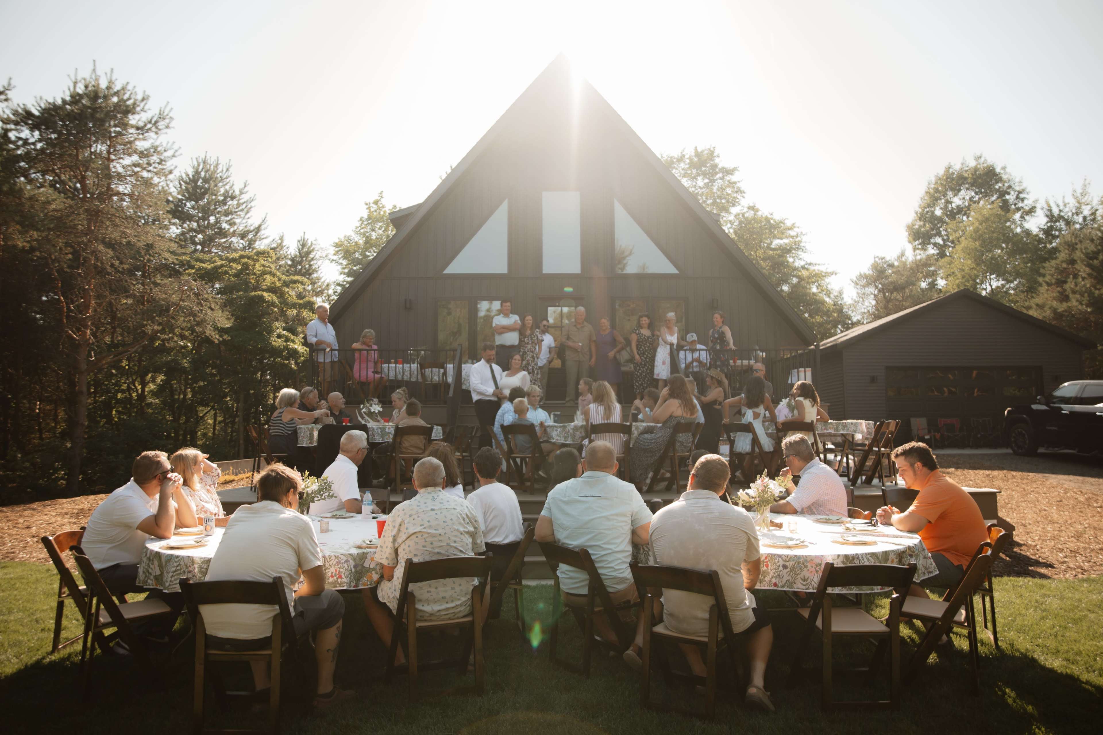 A large gathering takes place outdoors at a house, with numerous people seated at tables and others standing on a deck.