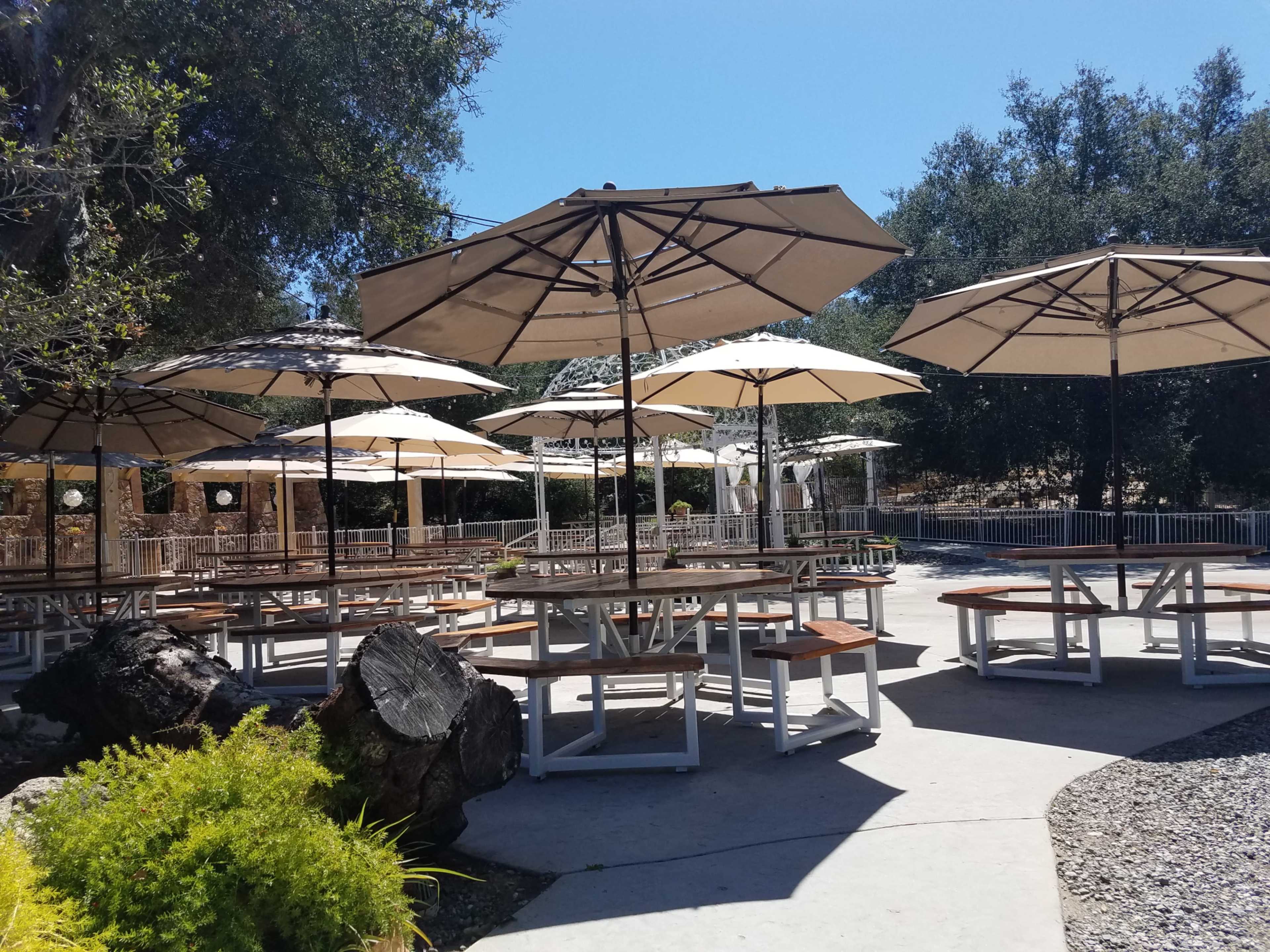 An outdoor seating area features several wooden tables with large umbrellas, surrounded by greenery and gravel paths.