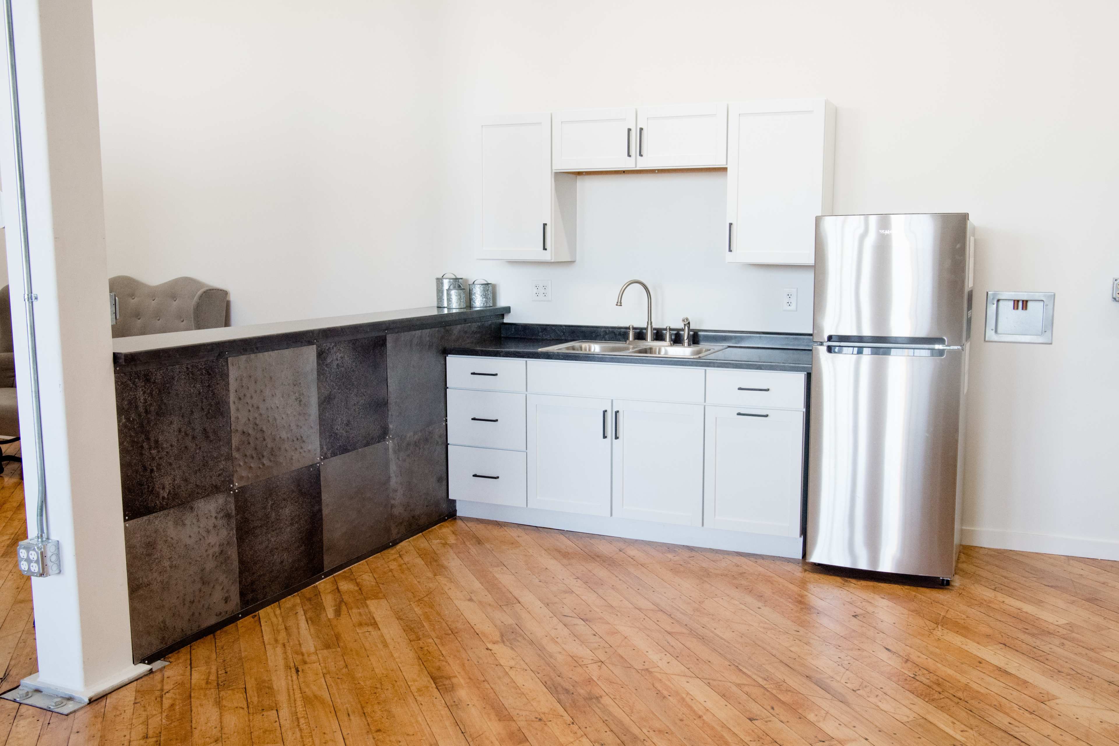 A modern kitchen with white cabinetry, a stainless steel refrigerator, and a countertop made of dark materials, set on a polished wooden floor.