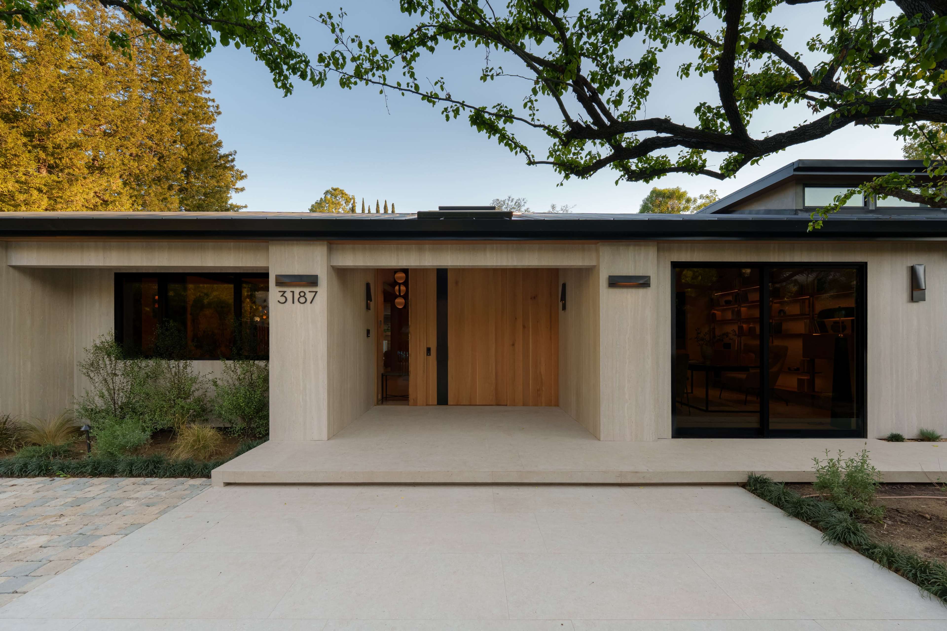 A modern house features a wooden entrance, large windows, and a stone pathway surrounded by greenery.