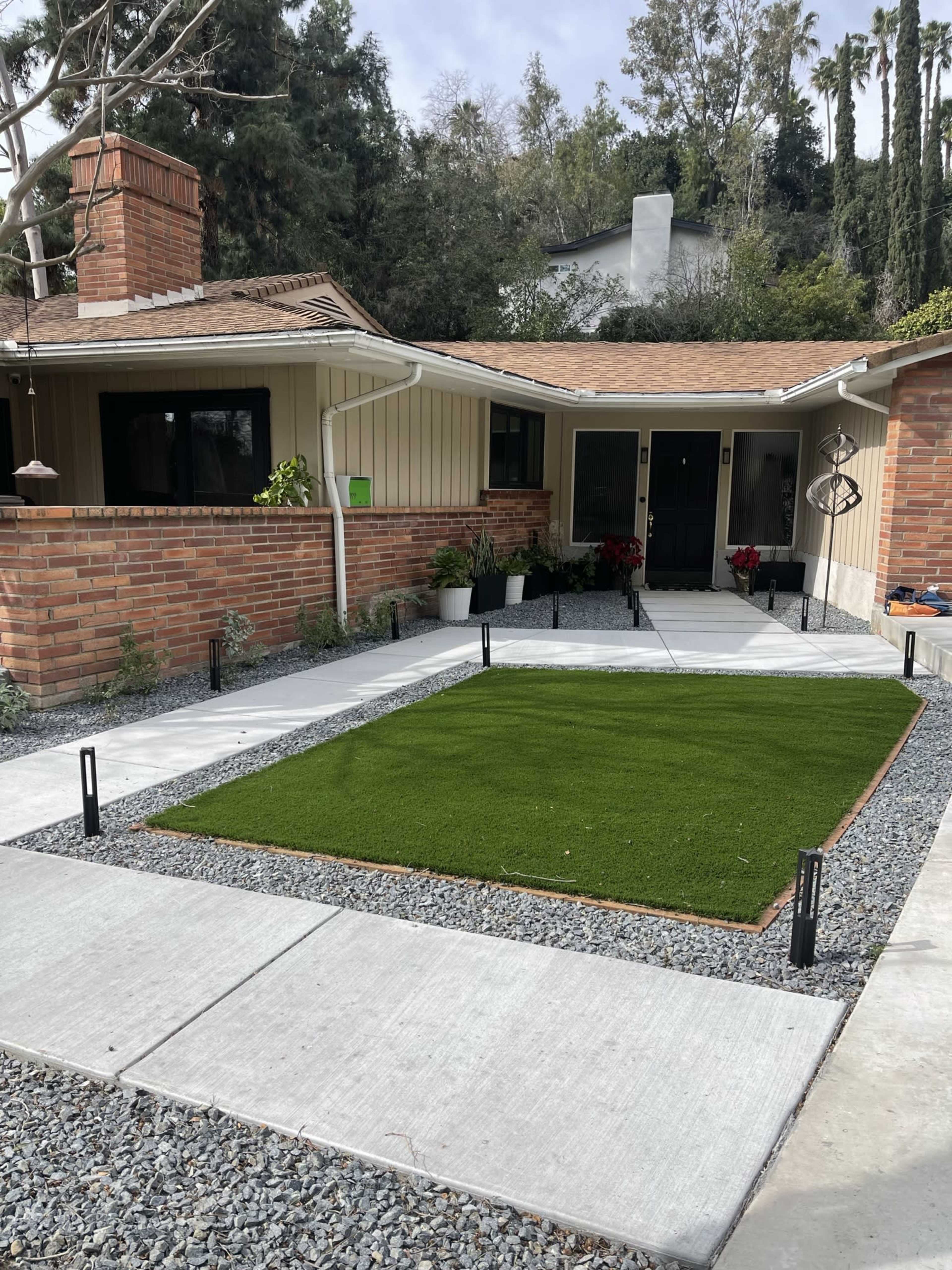 A landscaped front yard featuring a small patch of artificial grass surrounded by gravel and concrete walkways leading to a house.