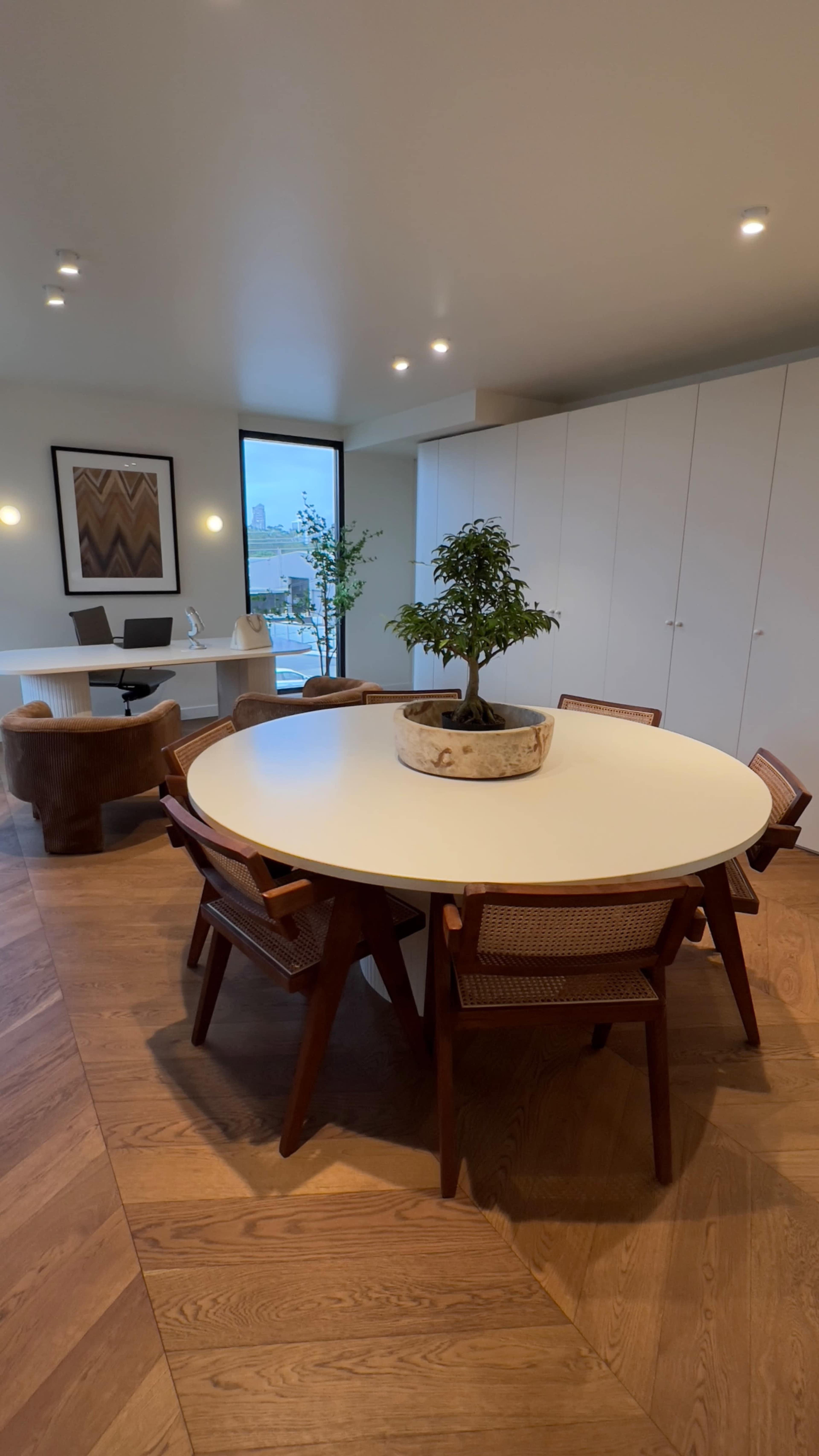 A round dining table surrounded by six wooden chairs sits in a spacious room with a large window and a stylish desk in the background.
