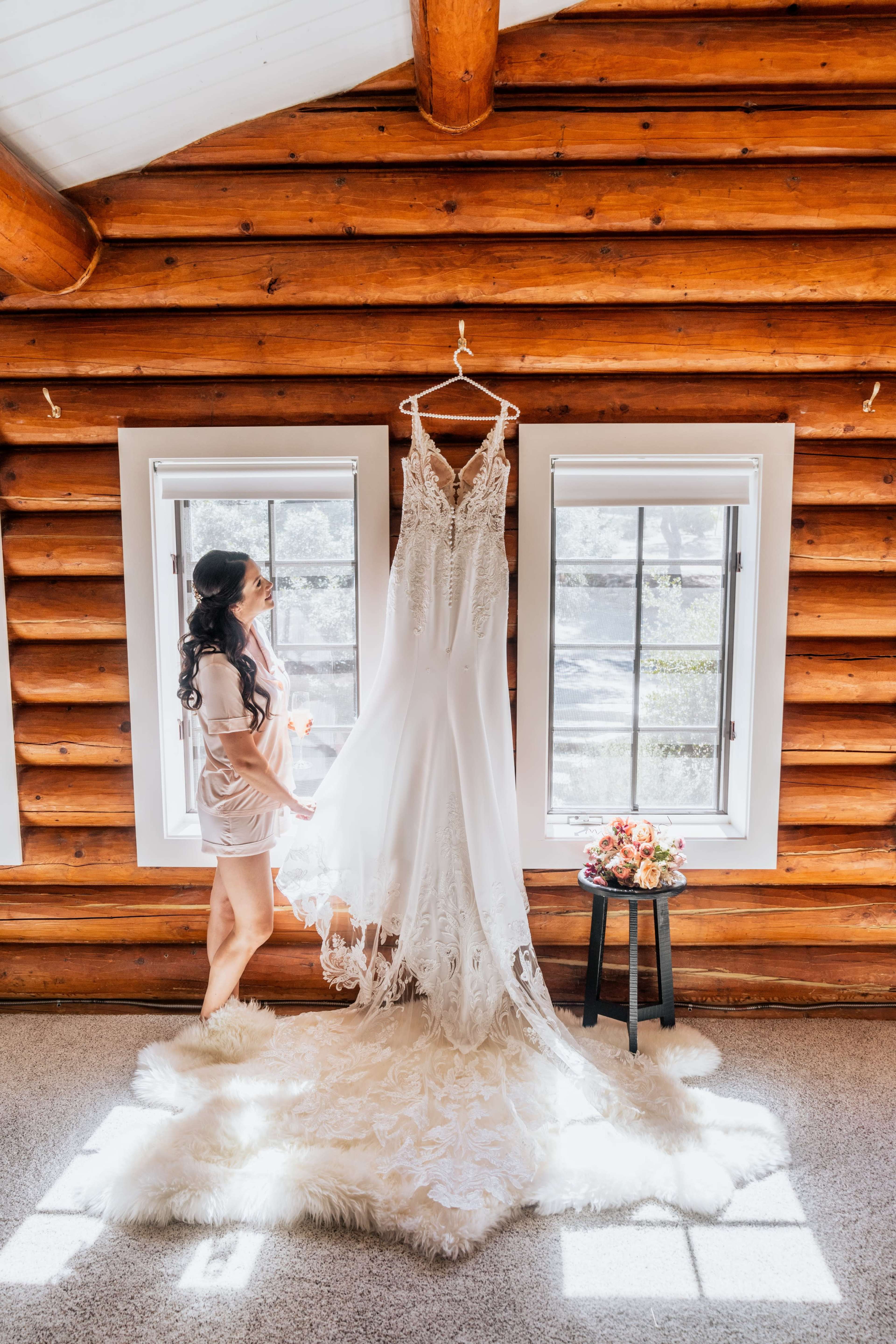 A woman stands beside a hanging wedding dress in a log cabin, with sunlight pouring through the windows.
